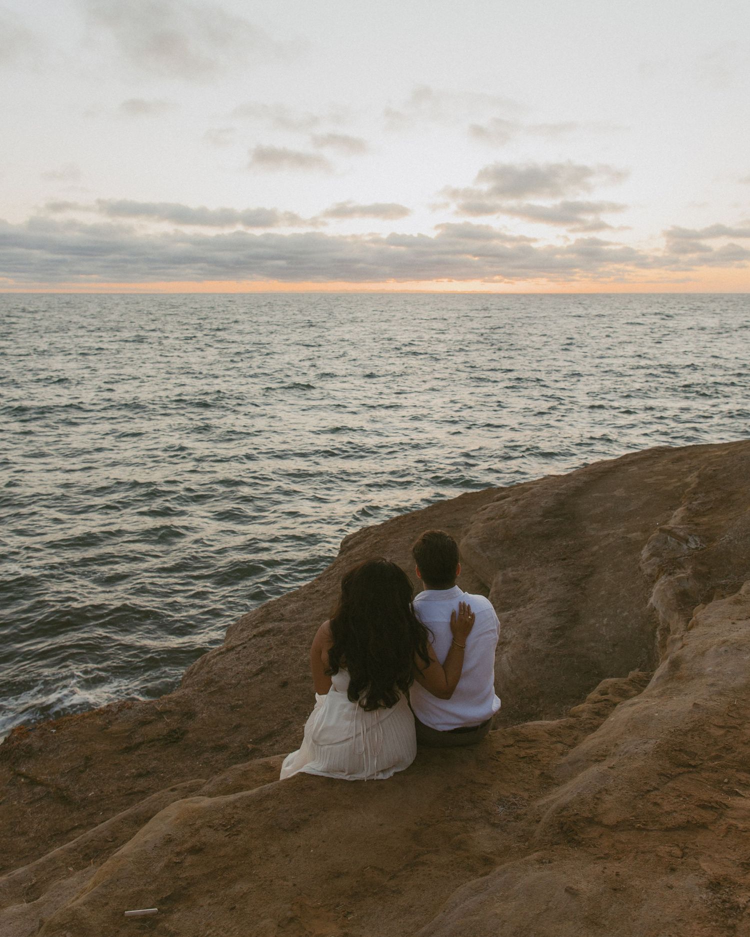 Couple sitting on rocky cliff overlooking calm ocean at sunset, viewed from behind.