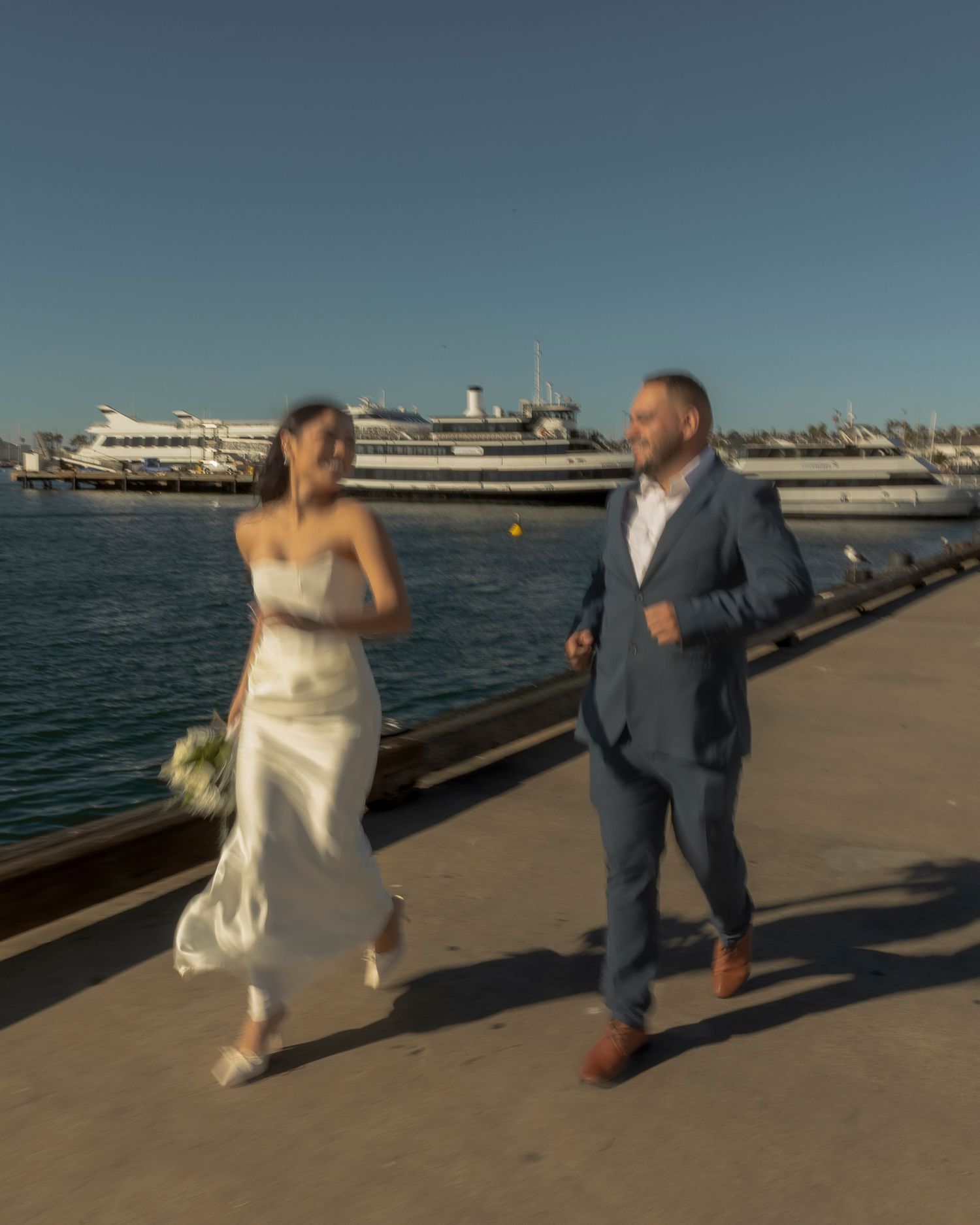 A couple in wedding attire runs joyfully along a waterfront pier with boats and a harbor in the background.