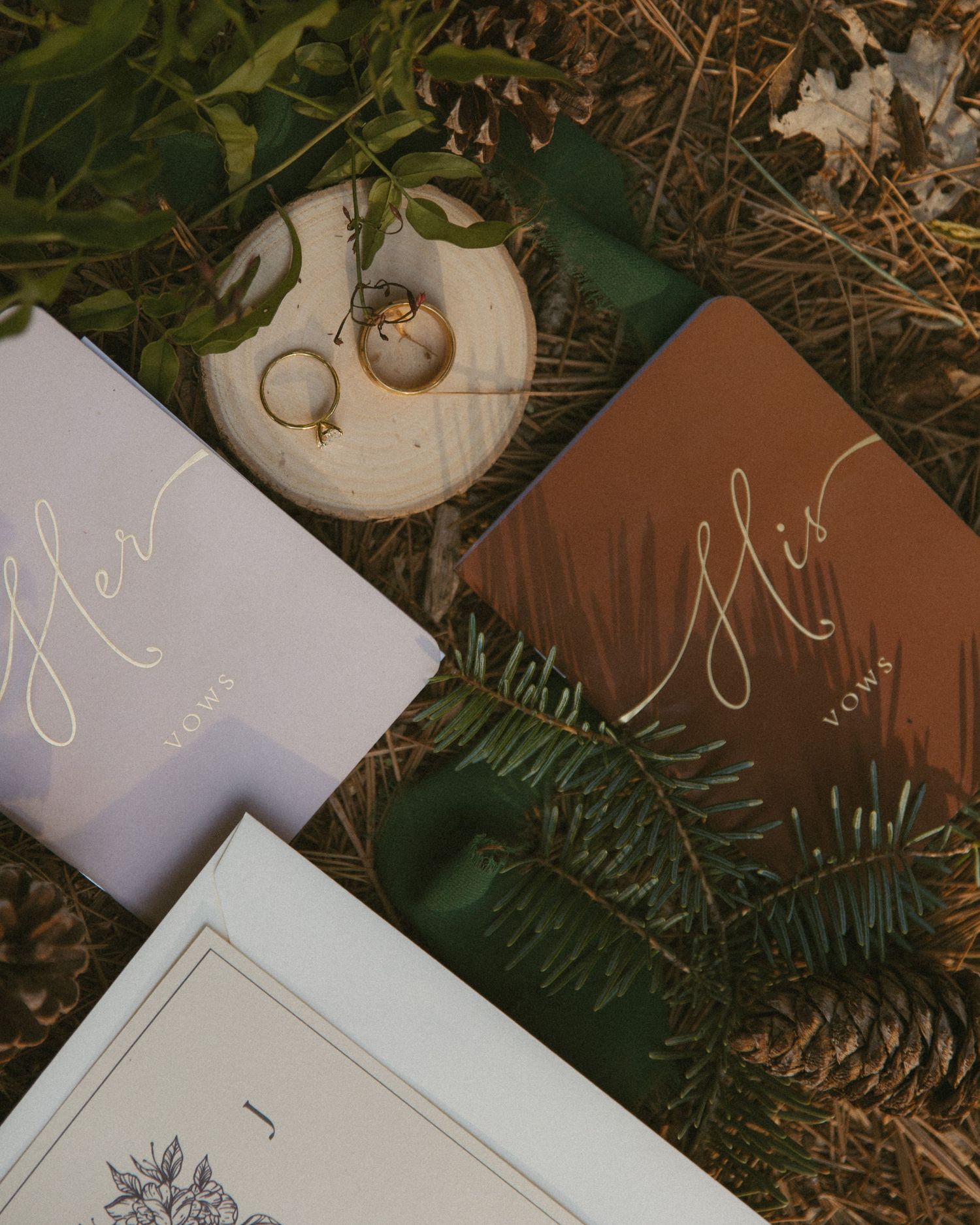 Her and His vow books with gold calligraphy, wedding rings on wood slice, pine branches, autumn wedding stationery flat lay.