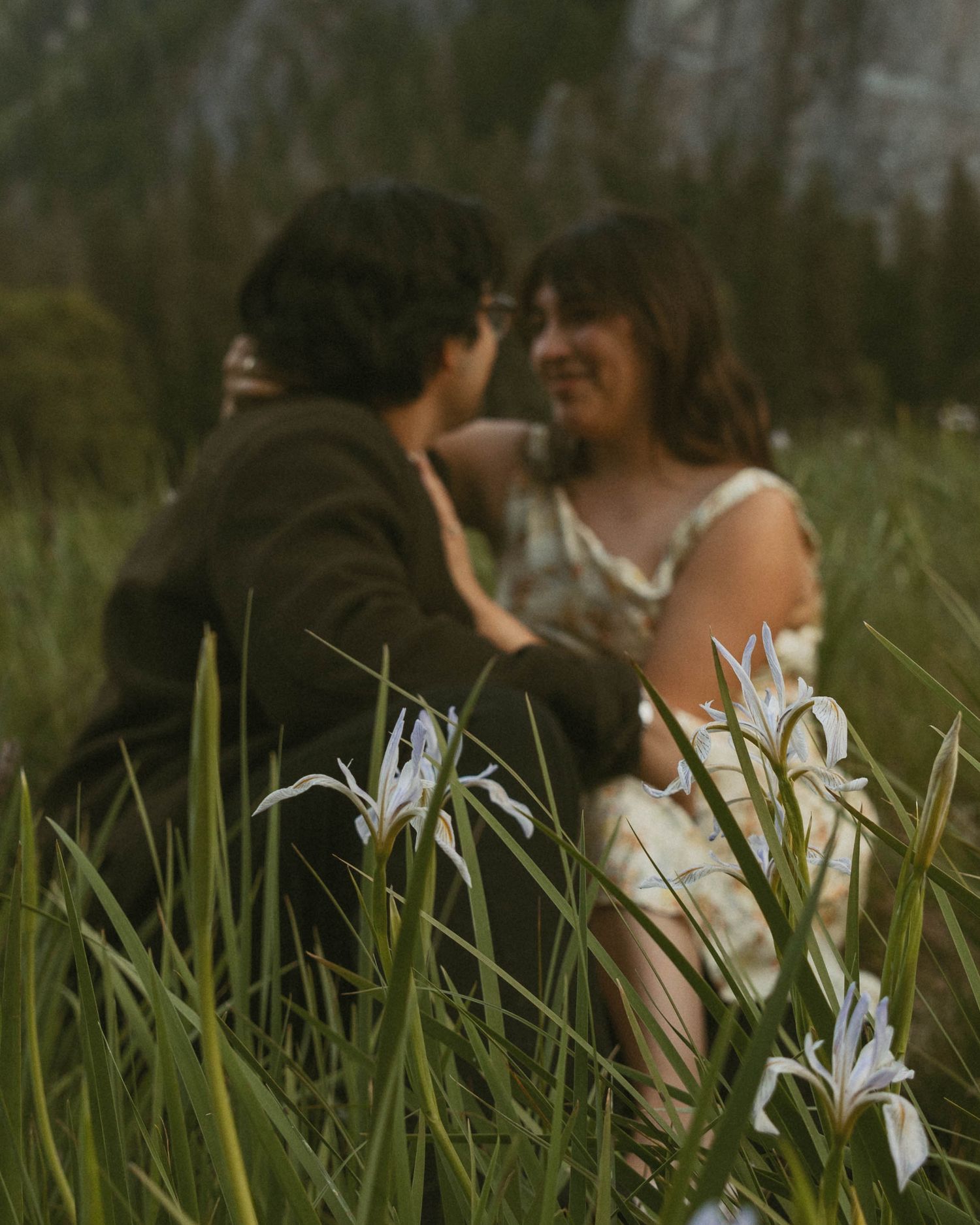 A couple sits together in a lush green field surrounded by white iris flowers in a romantic outdoor setting.