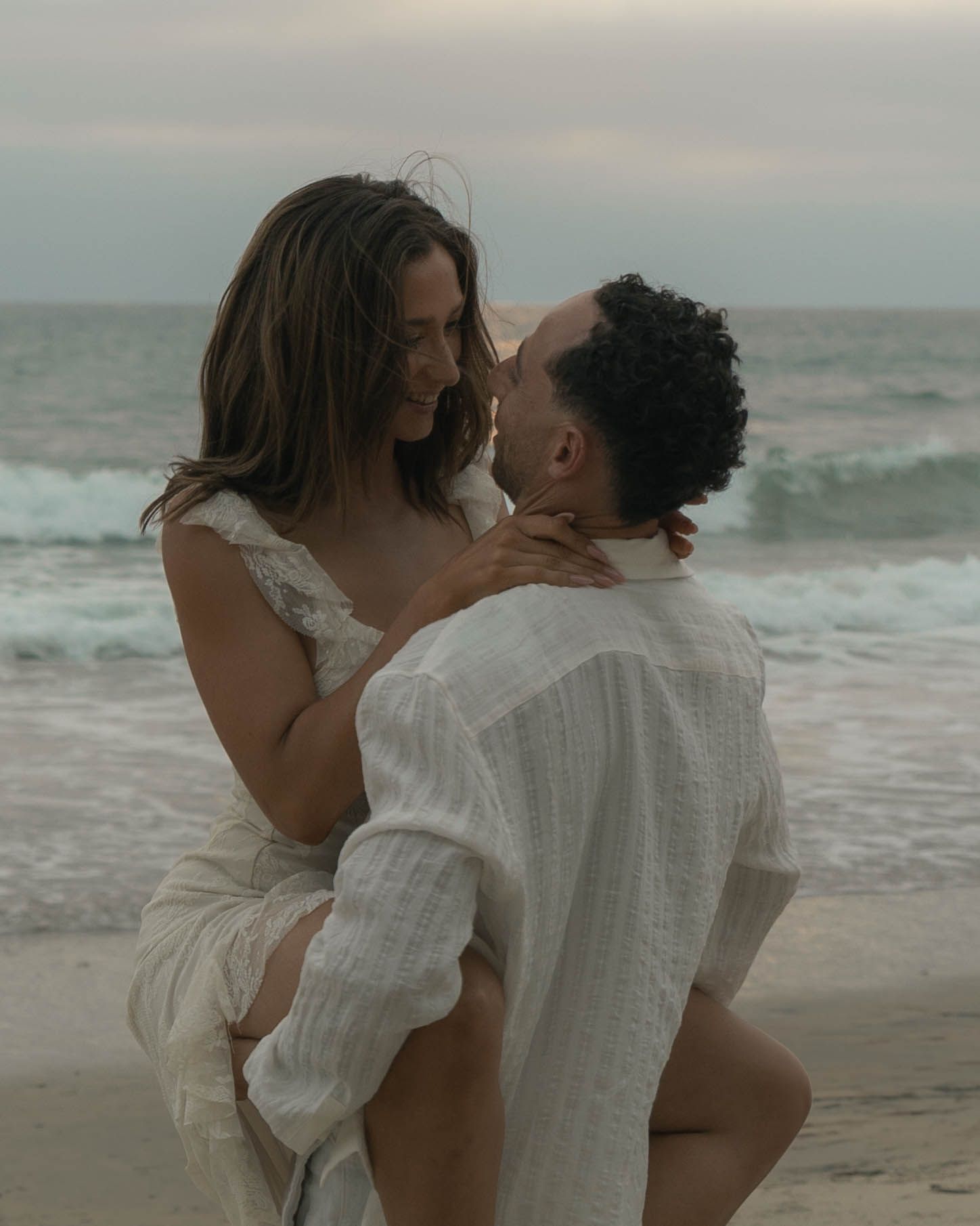 A couple in white clothing shares tender moments by the ocean waves.