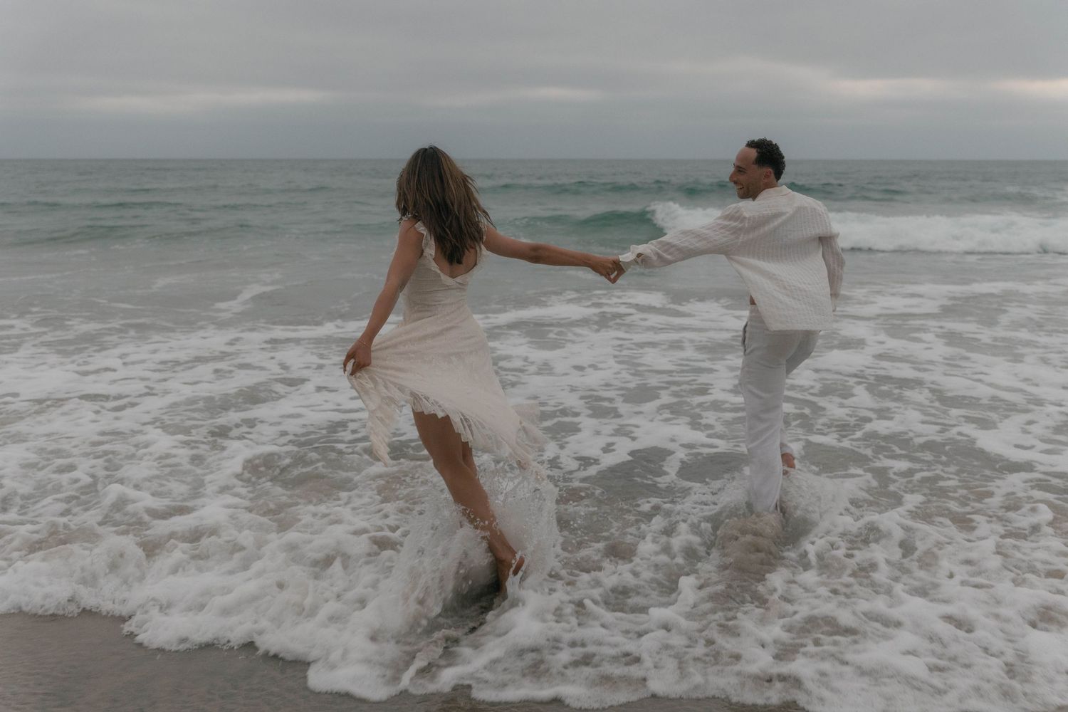 A couple plays joyfully in the ocean waves at the beach during sunset.