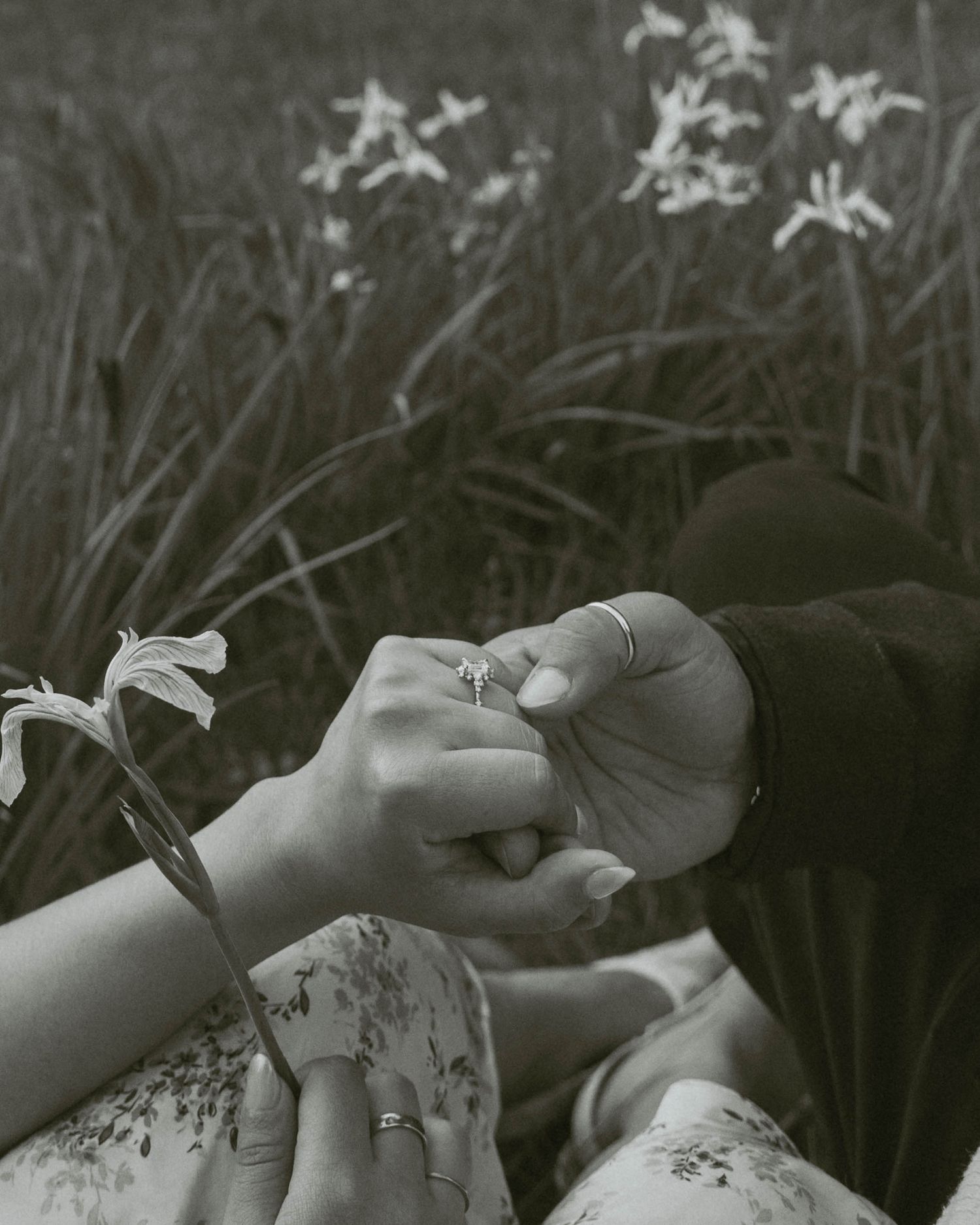 Black and white photo of two people holding hands with a ring, surrounded by blooming flowers in a field.