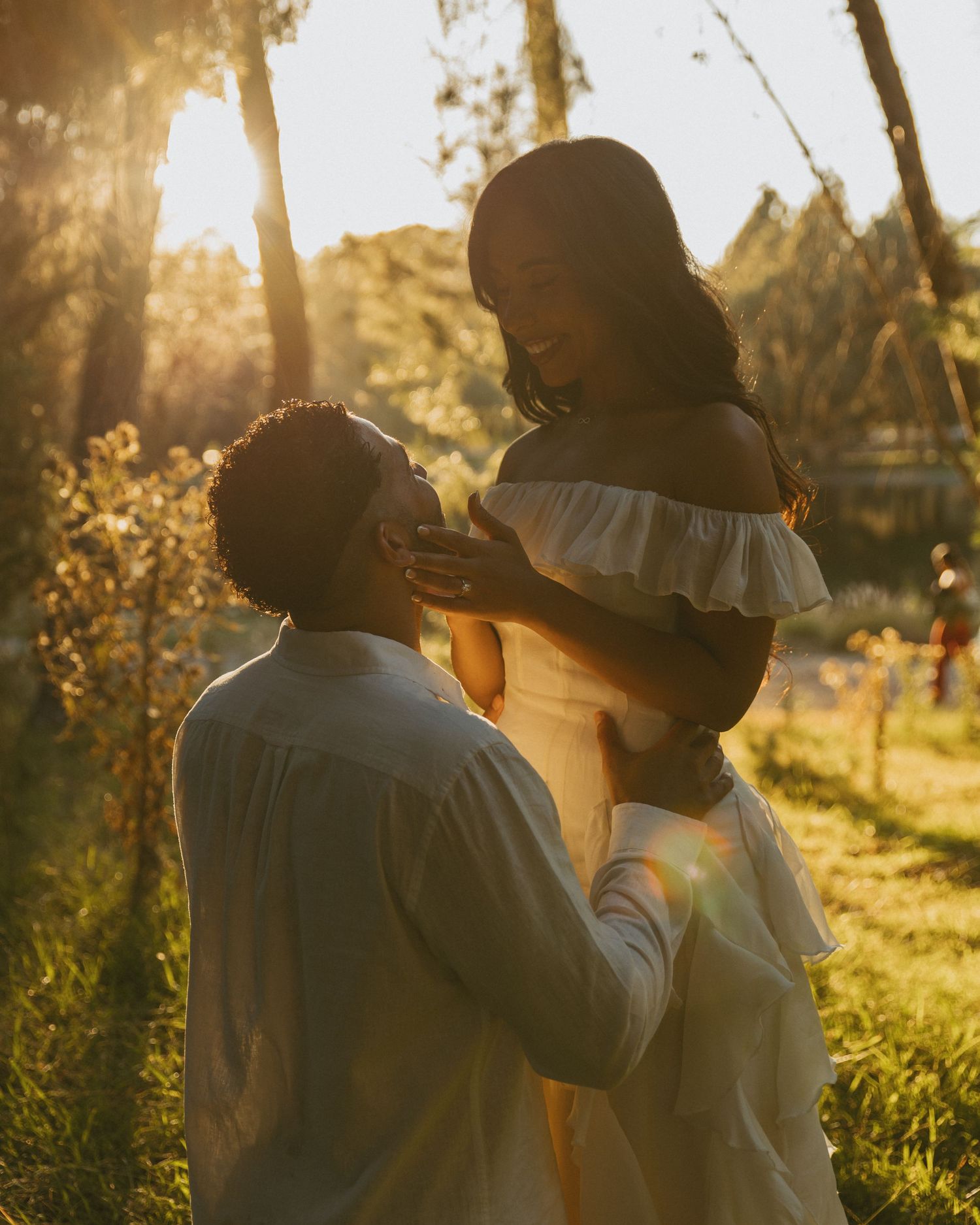 Couple sharing a romantic moment in golden sunset light among trees, woman in white off-shoulder dress.