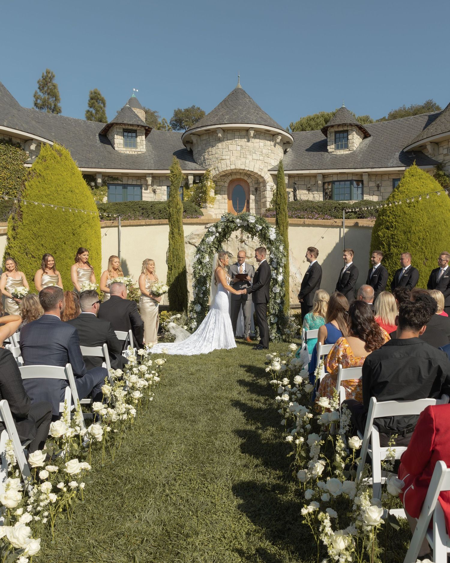 Outdoor wedding ceremony setup with white chairs and floral arrangements in front of an elegant estate.