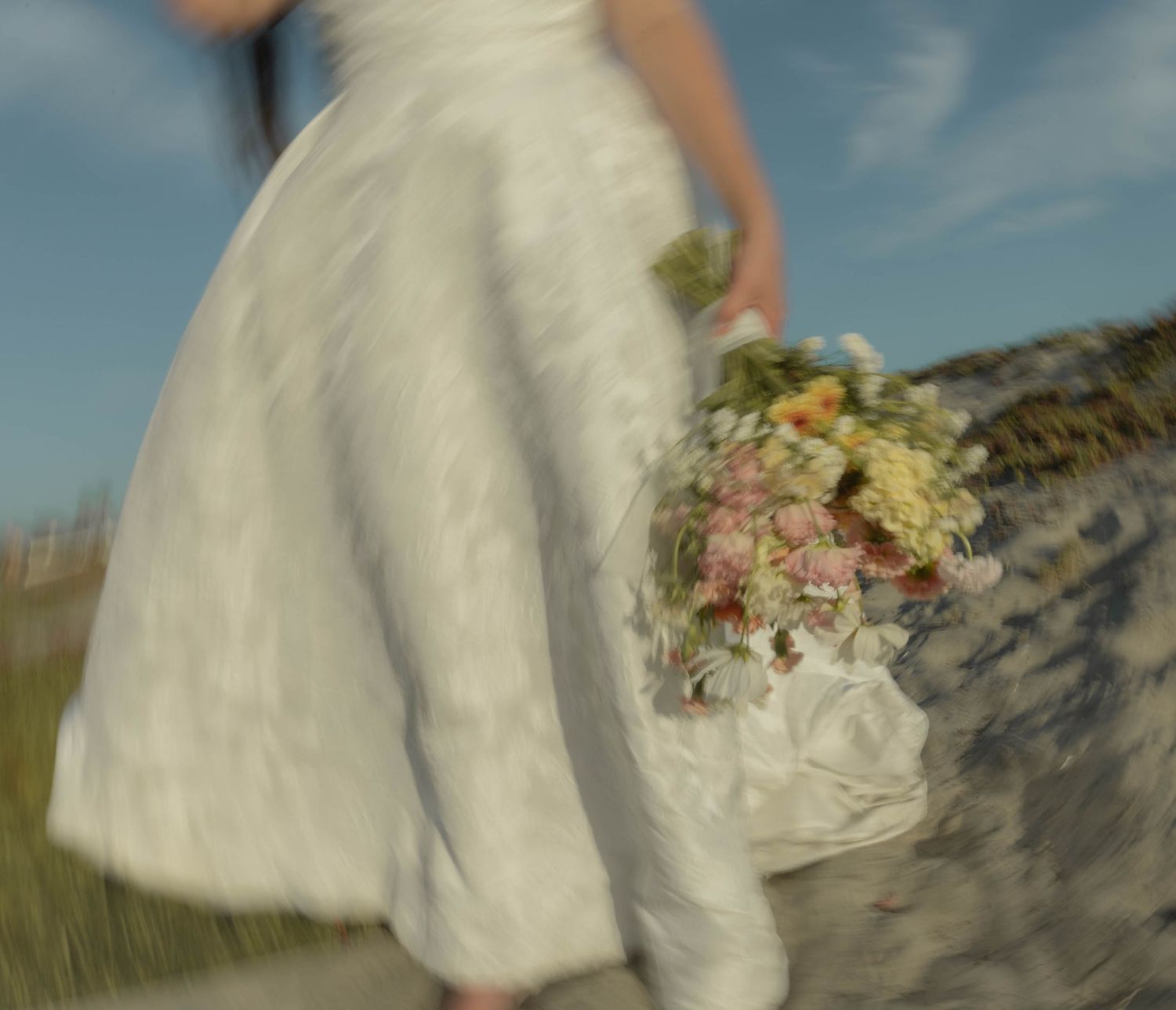 A flowing white wedding dress catches the breeze on a sunny beach.