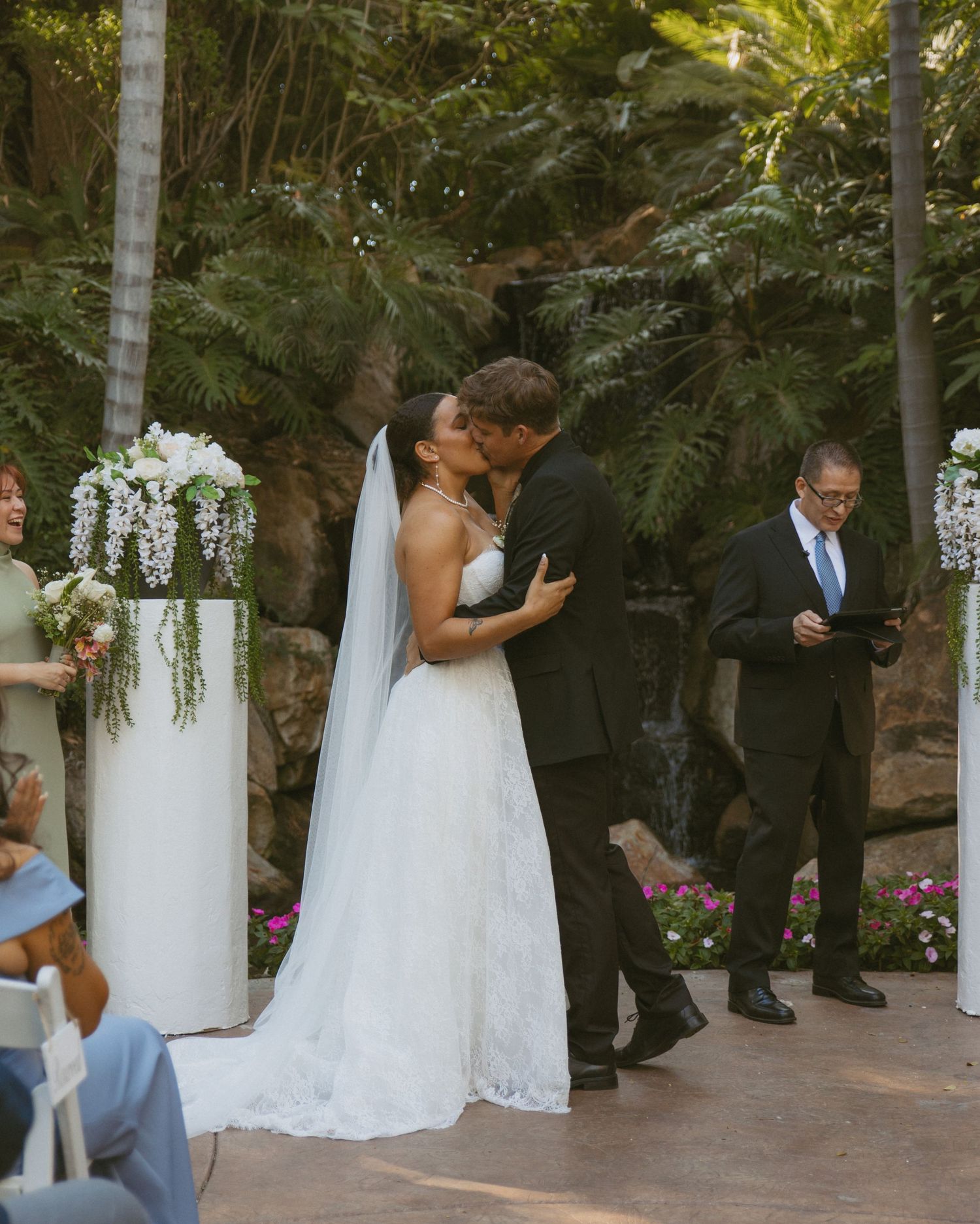 Bride and groom share first kiss at outdoor garden wedding ceremony with white floral arrangements.