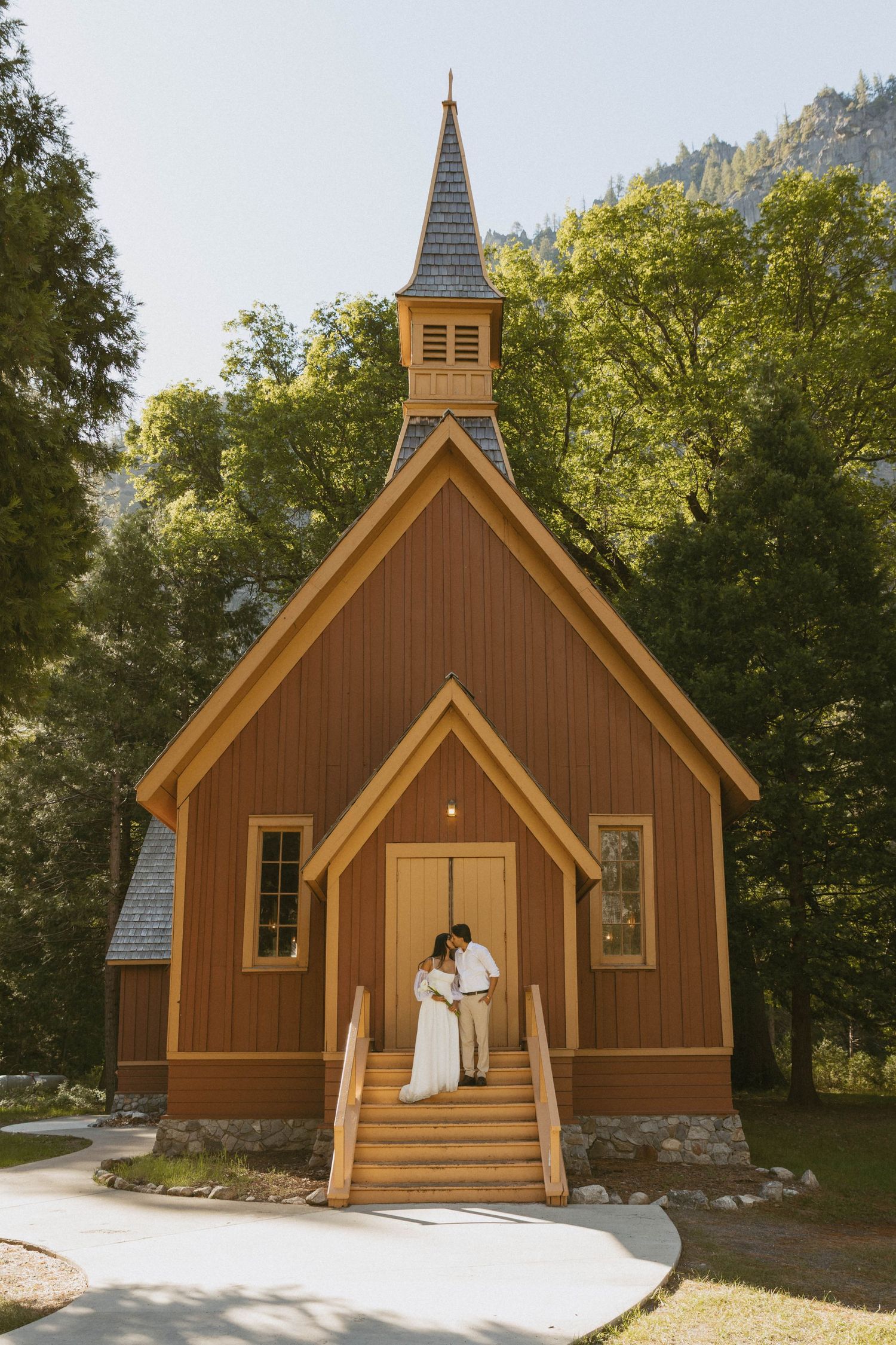 Couple embracing on steps of rustic wooden chapel surrounded by lush trees in Yosemite Valley wedding photo.