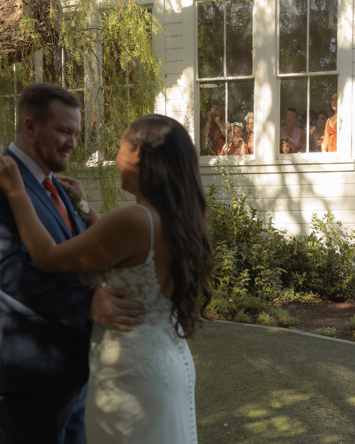 Bride and groom share first dance outdoors while wedding guests watch through a white building window.