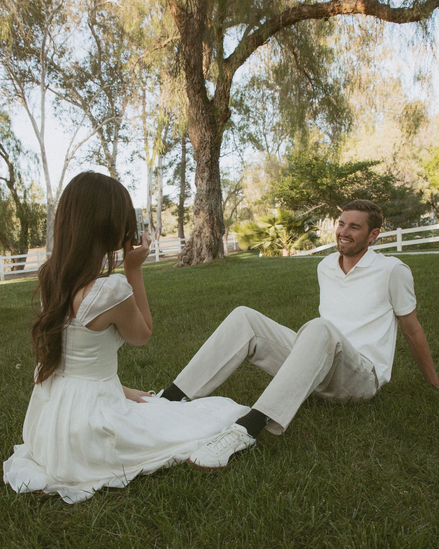 A couple in white outfits sits on green grass near trees, with the woman photographing the smiling man.