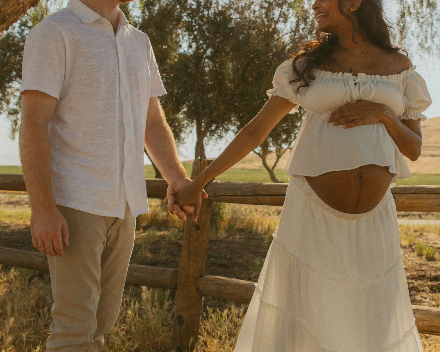 Couple holding hands outdoors at golden hour, pregnant woman in white dress cradling baby bump near rustic fence.