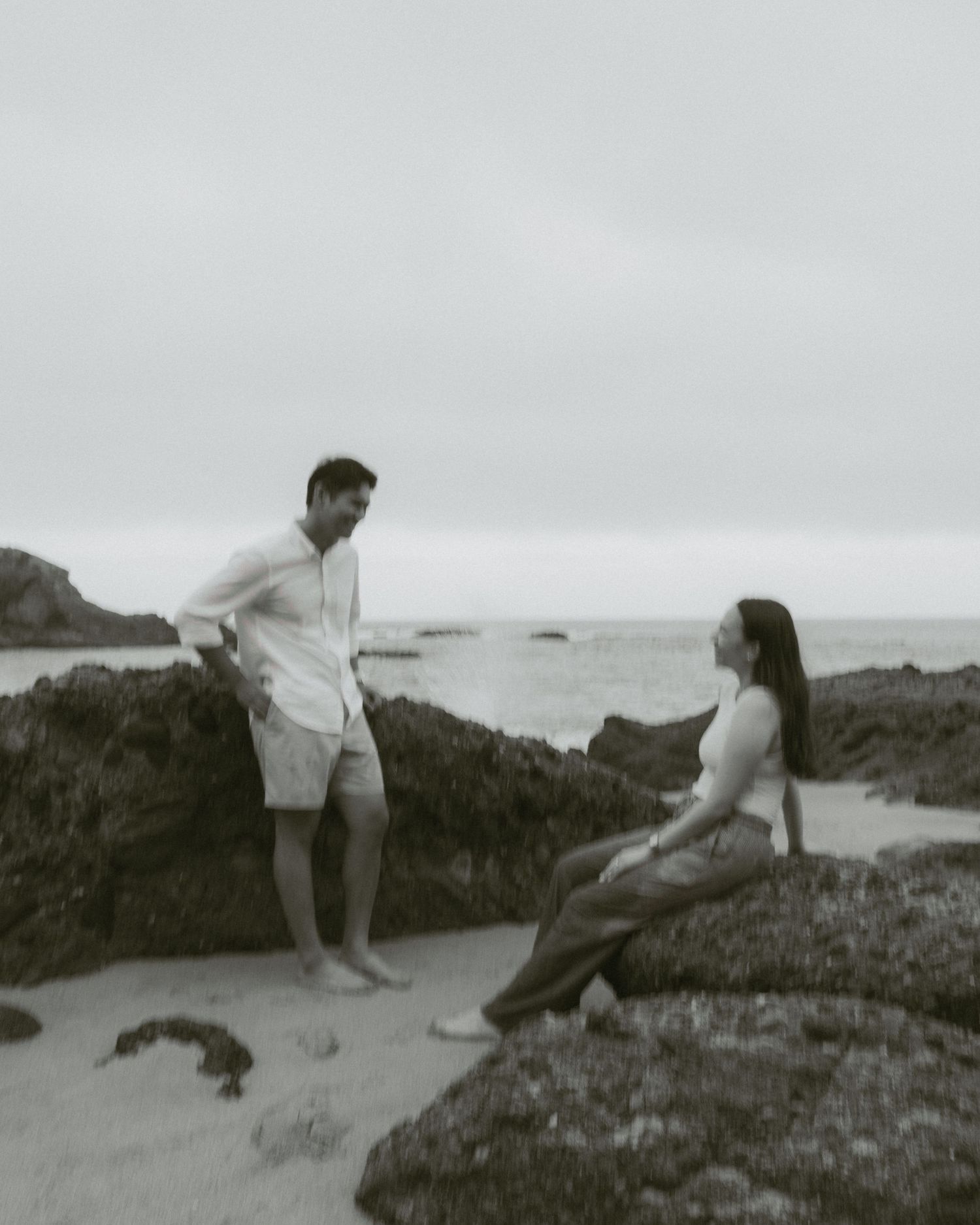 Black and white photo of a couple conversing on a rocky beach with ocean waves in the background.