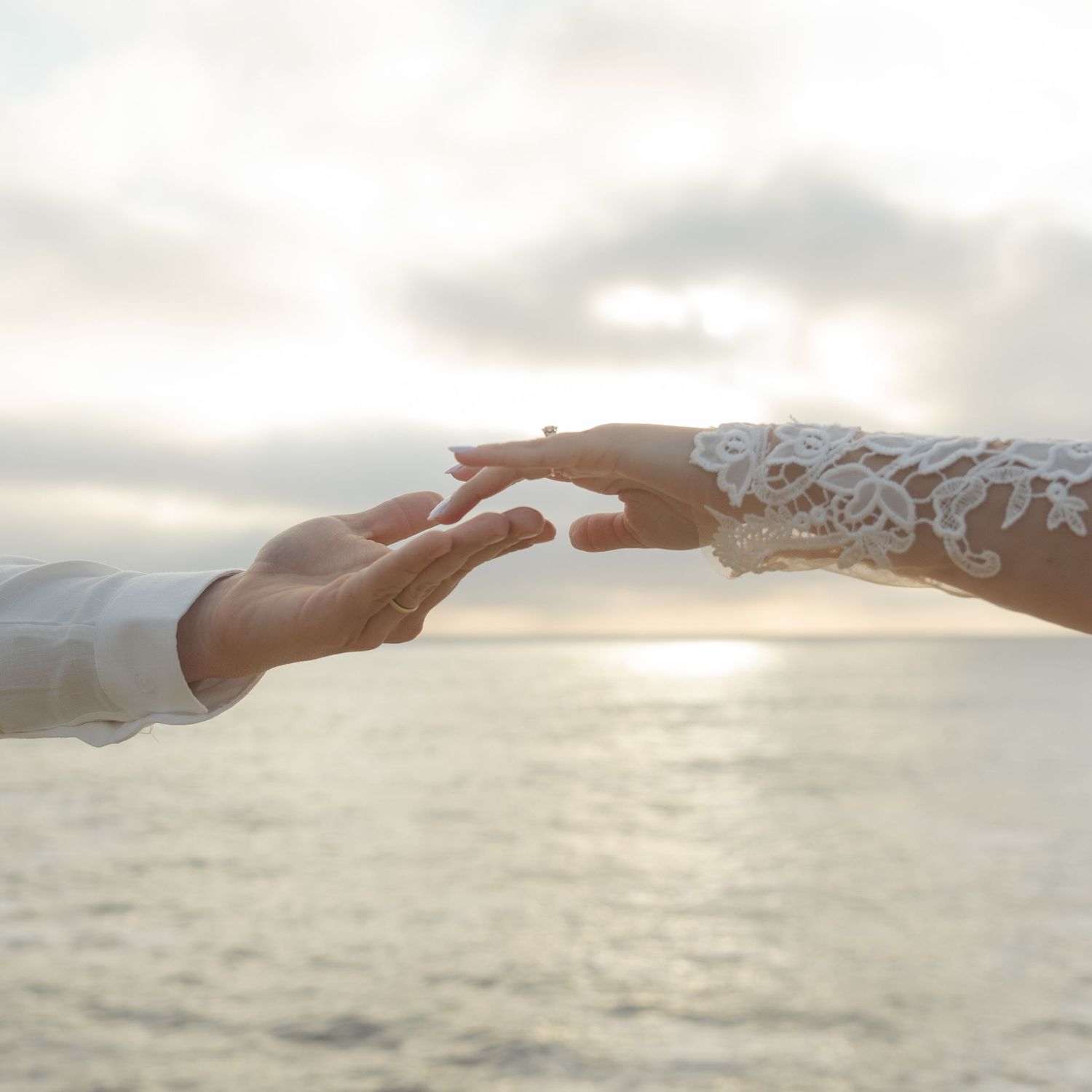 Two hands reaching towards each other against a sunset sky, with one wearing delicate white lace from a wedding dress sleeve.