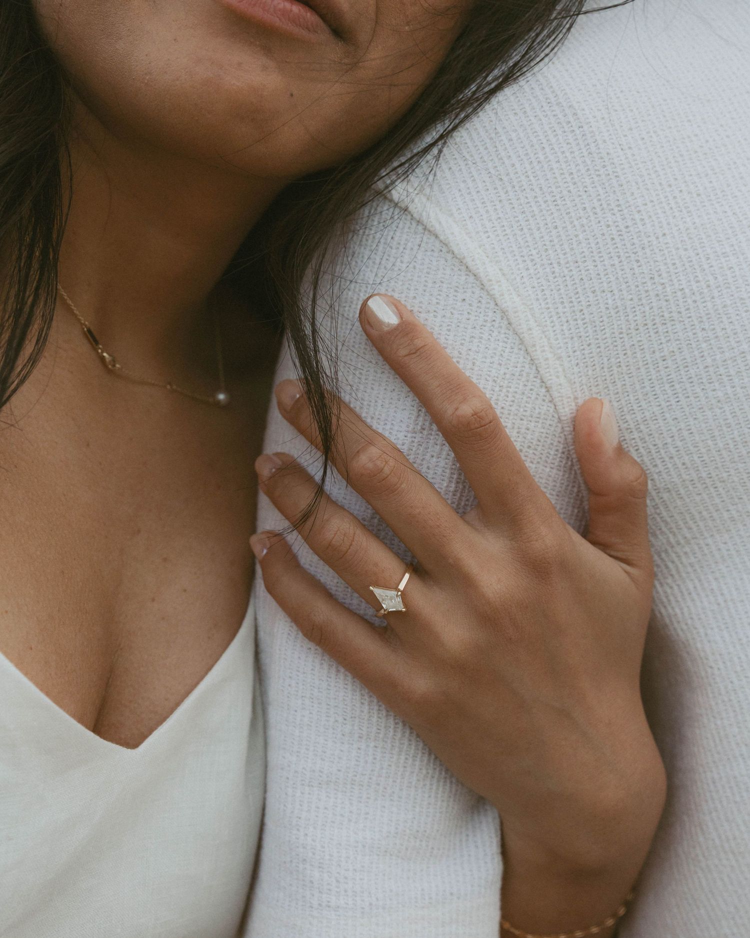 Woman wearing a gold diamond engagement ring and delicate necklace, resting hand on partner's white sweater.