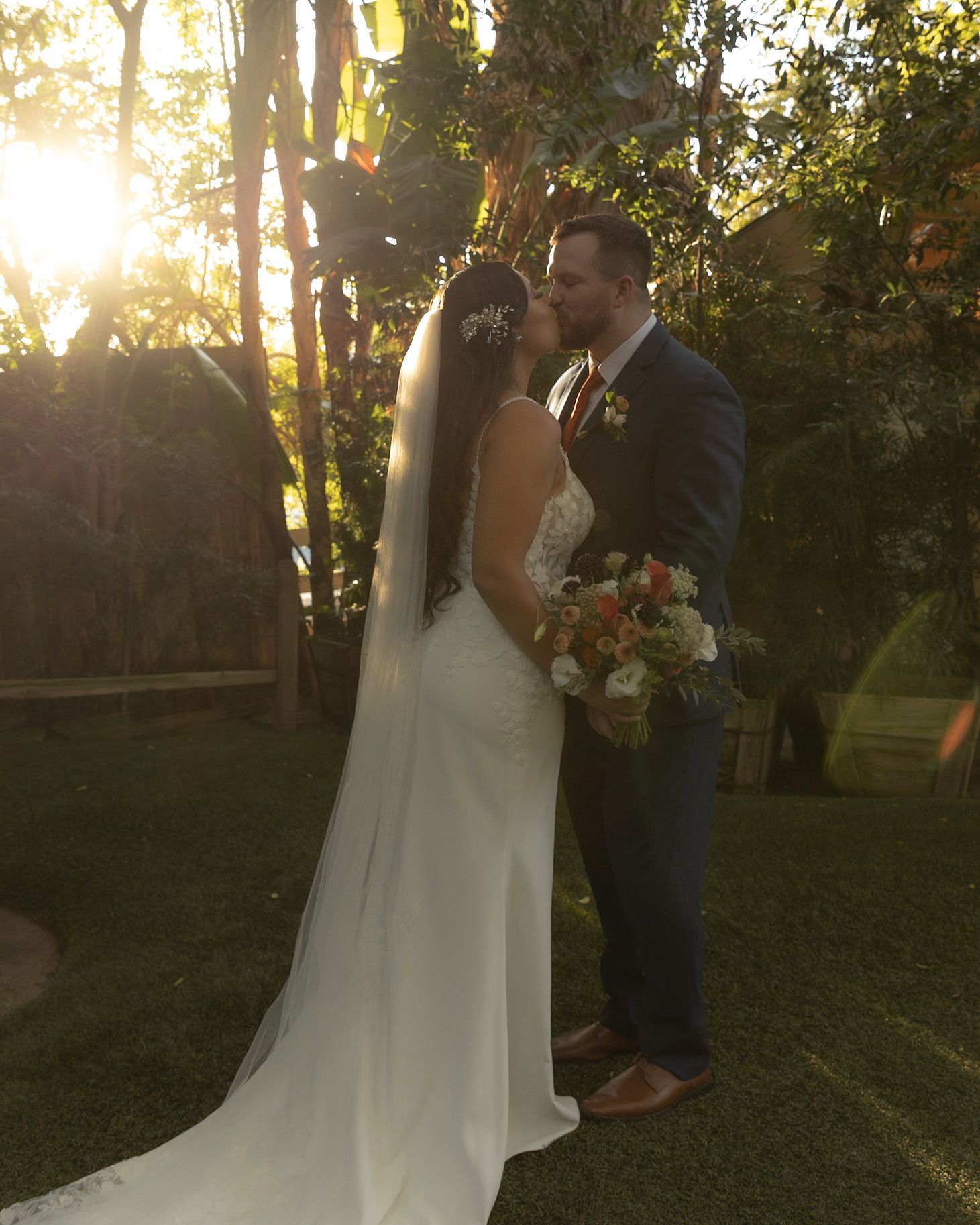 Bride and groom share a romantic sunset kiss outdoors, bride in white gown with veil holding colorful bouquet.