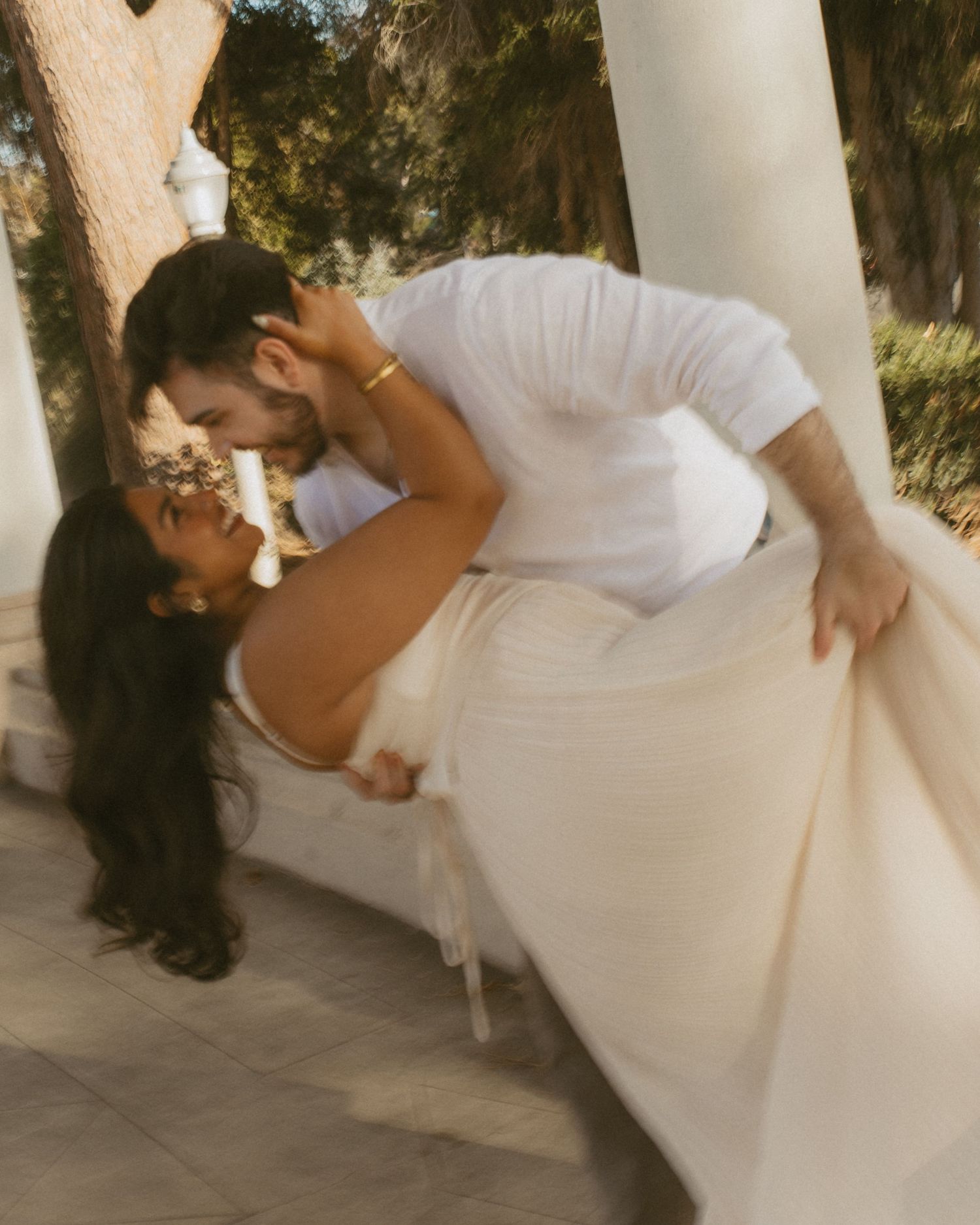 A couple shares a romantic dip kiss outdoors, dressed in white near elegant columns surrounded by lush greenery.