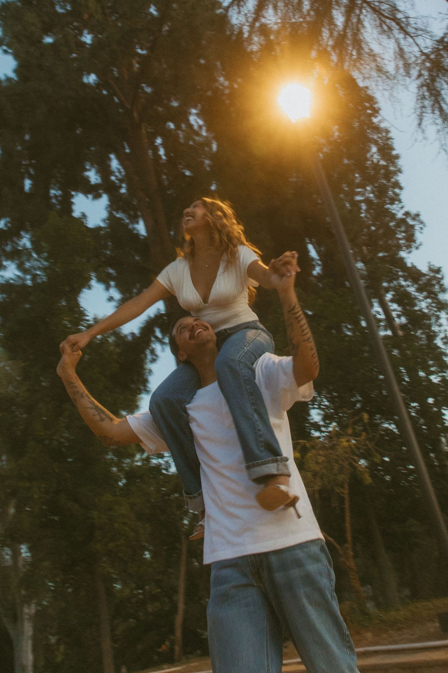 A woman sits on a man's shoulders in a sunlit park, both laughing joyfully among tall trees at golden hour.