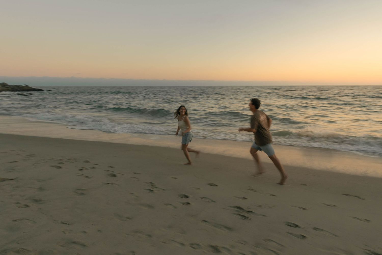 Two people run playfully along the shoreline at sunset with waves crashing nearby.