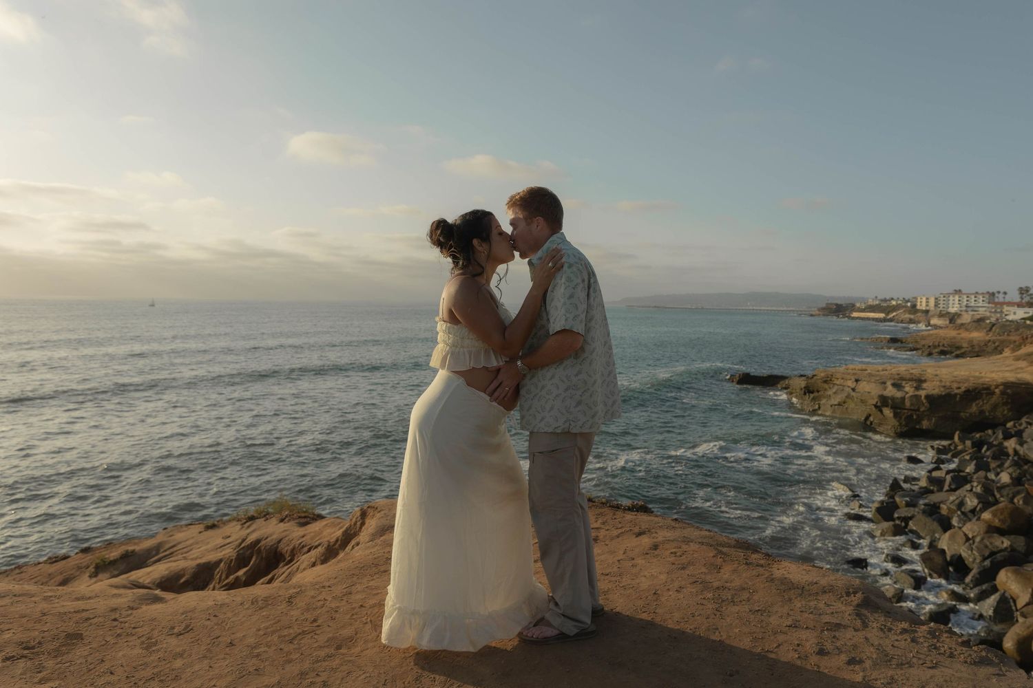 A couple sits together on coastal rocks overlooking the ocean at sunset.