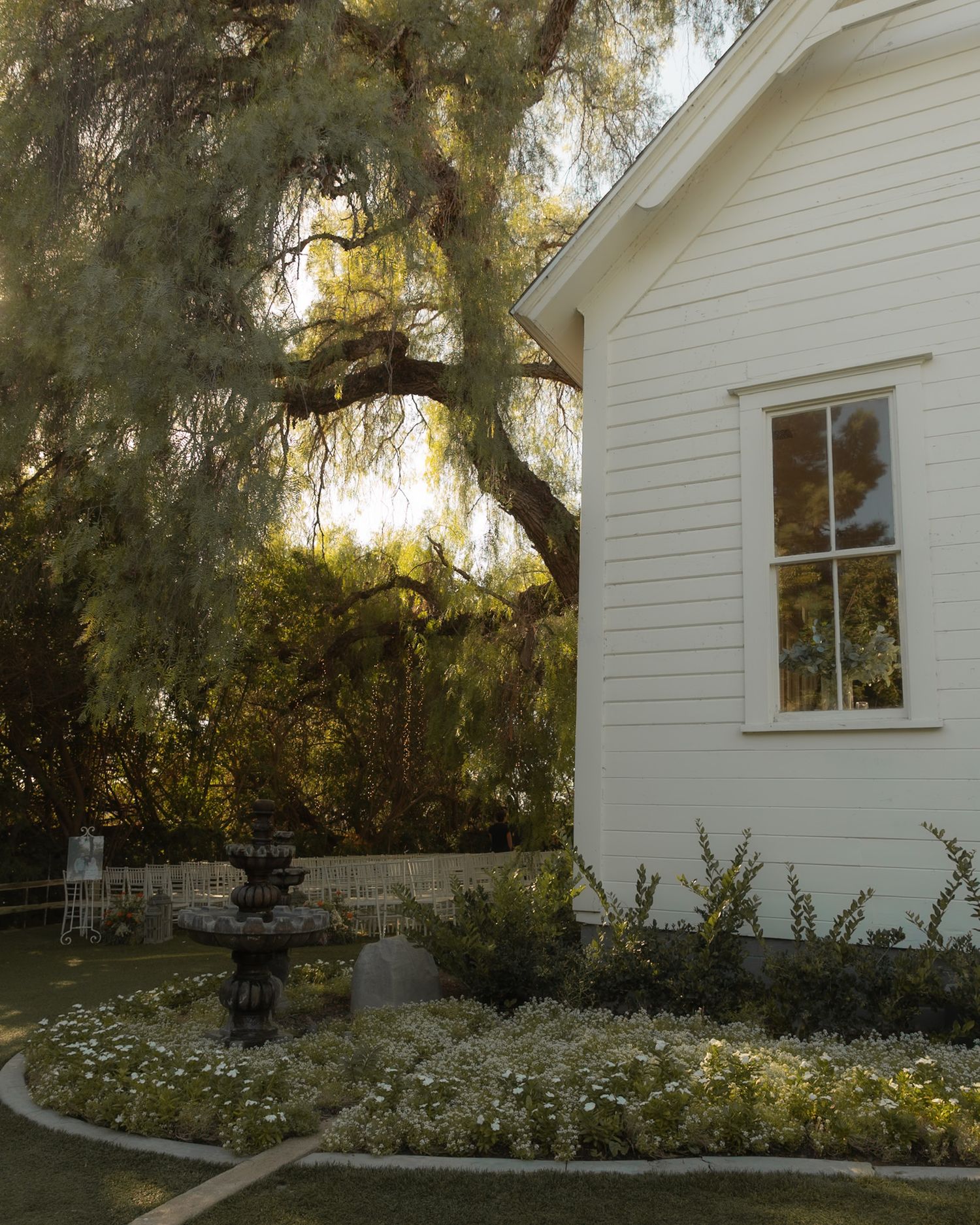 Charming white chapel with a stone fountain surrounded by white flowers and a weeping willow tree.
