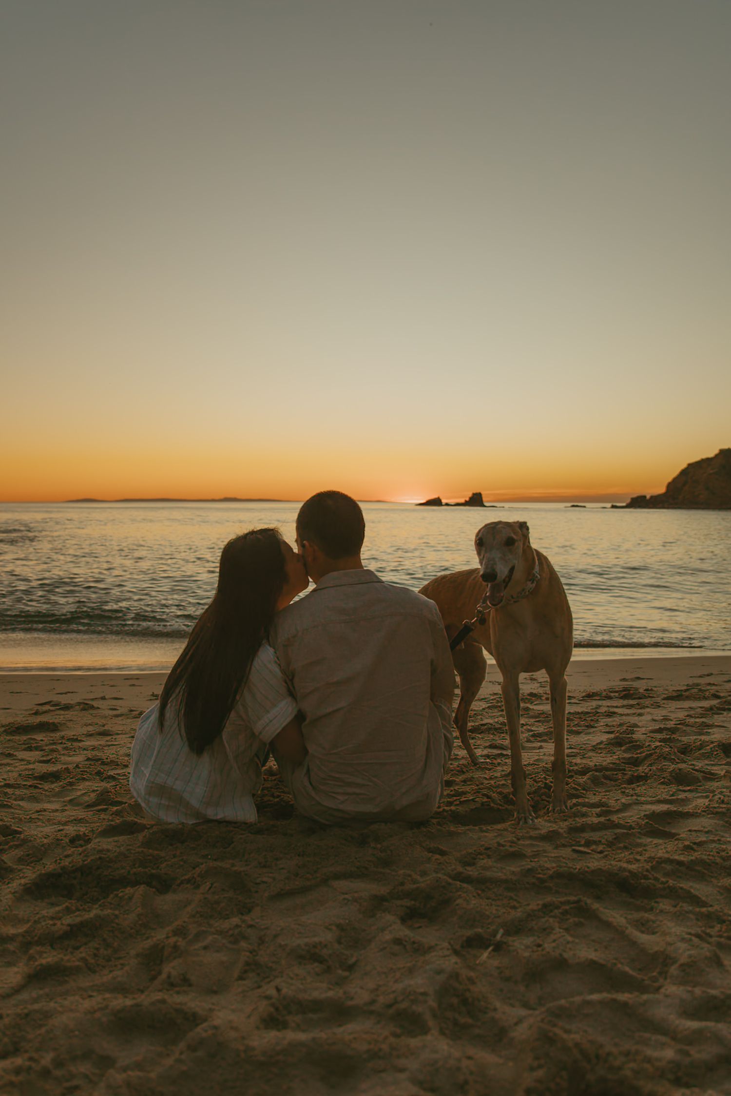 Couple sitting on beach sand with their dog watching a golden sunset over the ocean.