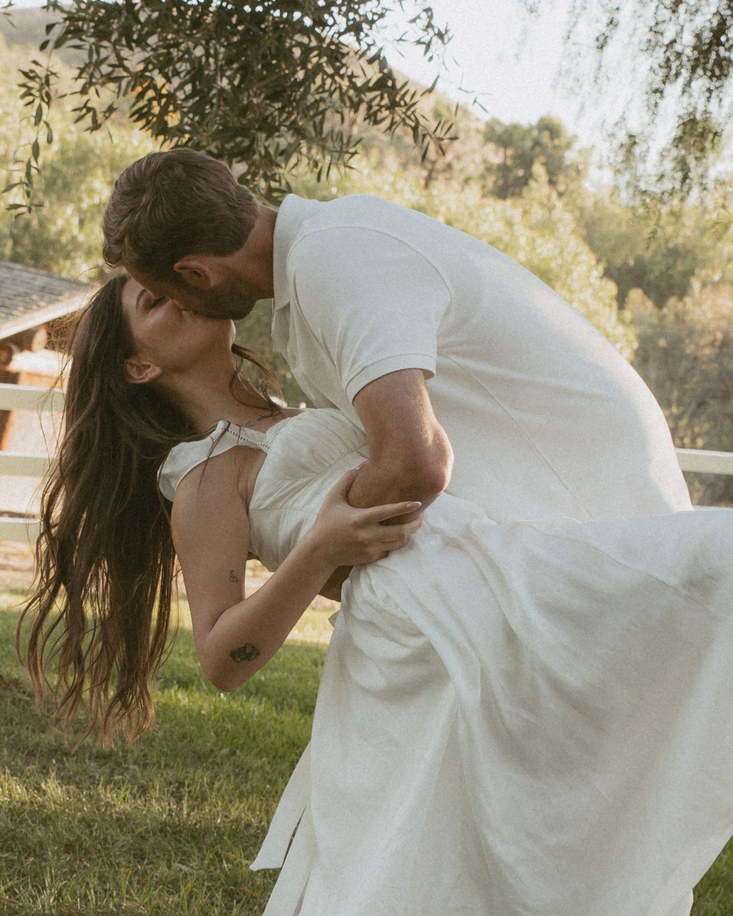 A couple shares a romantic dip kiss outdoors, the woman in a white dress, surrounded by lush greenery.