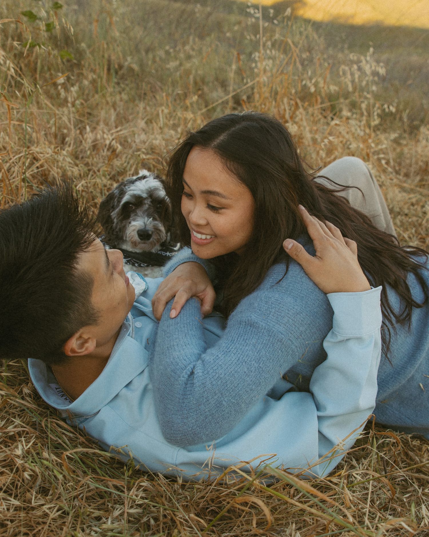 Couple lying in golden grass field smiling at each other with a small black and white dog between them.