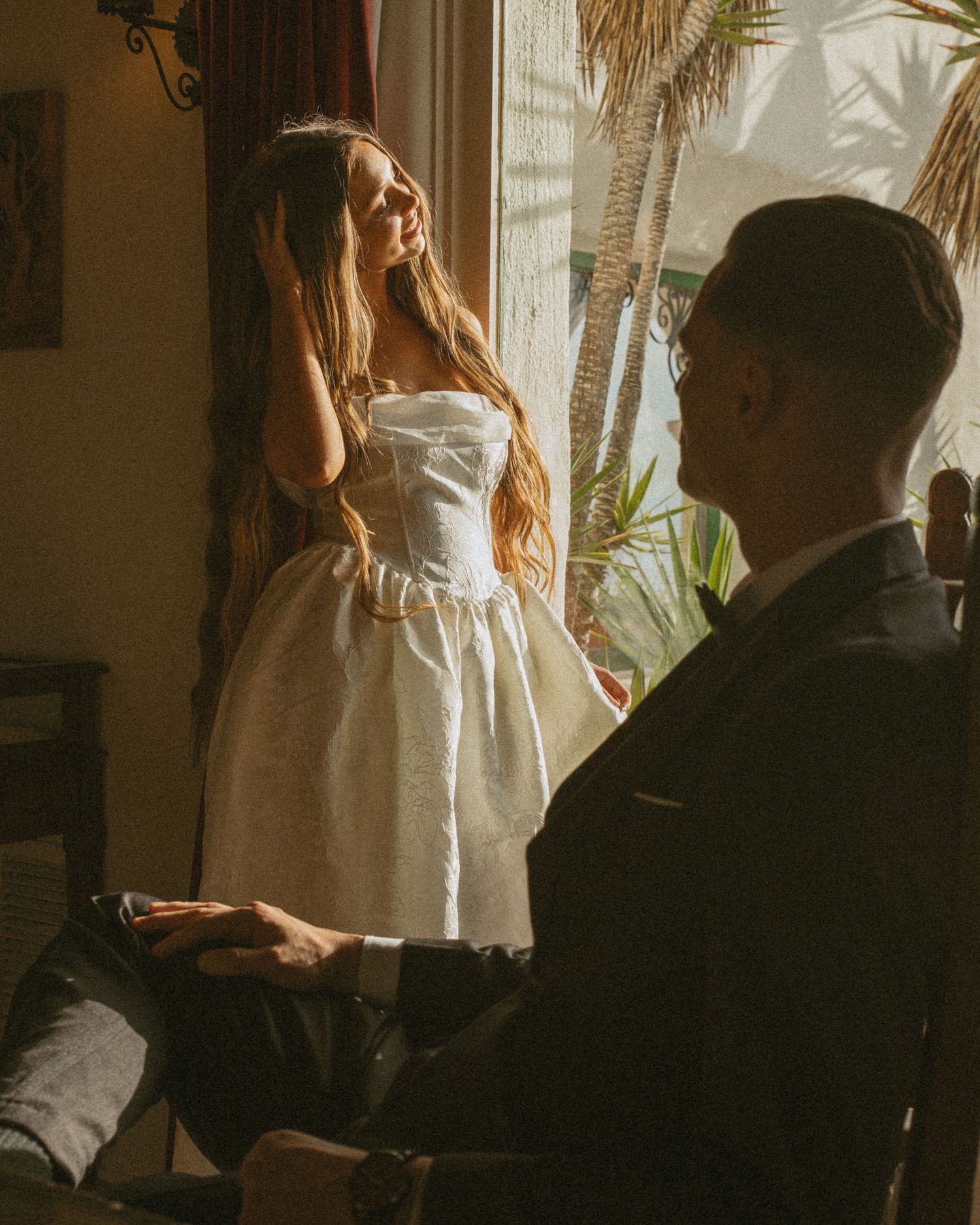 A bride in a white strapless mini dress holds hands with a groom in a dark suit near a sunlit window.