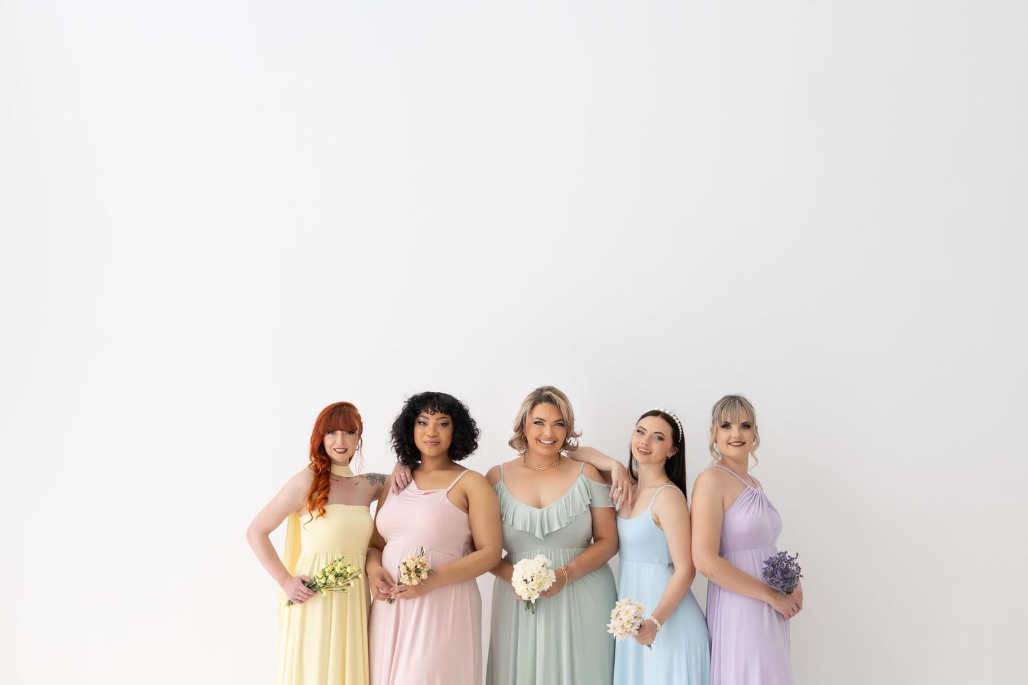 A group of people dressed in pastel bridesmaid dresses and gowns stand together in a lineup against a white backdrop.