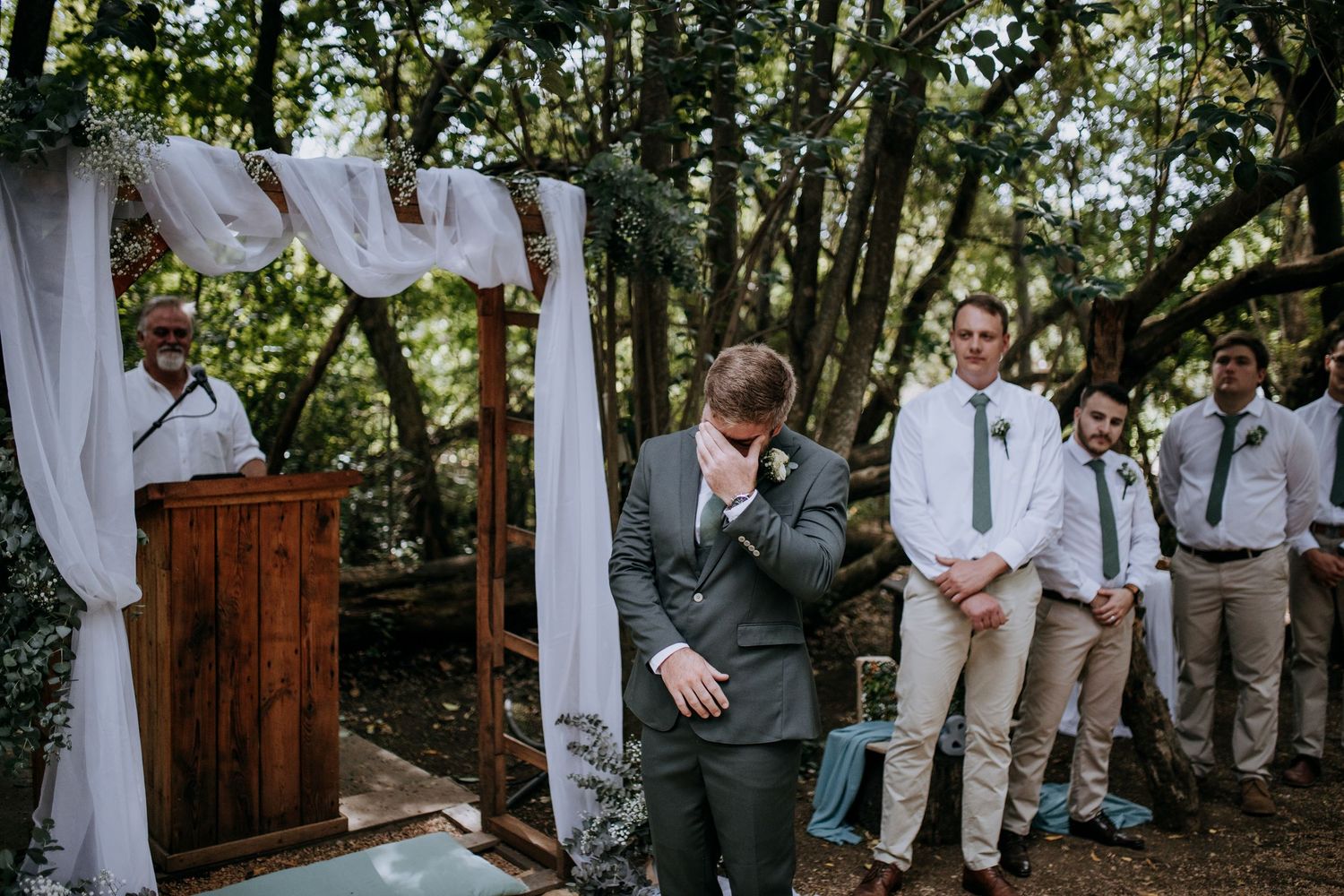 Members of a wedding party in gray suits and khakis walk through a wooded outdoor setting with sheer white drapes.