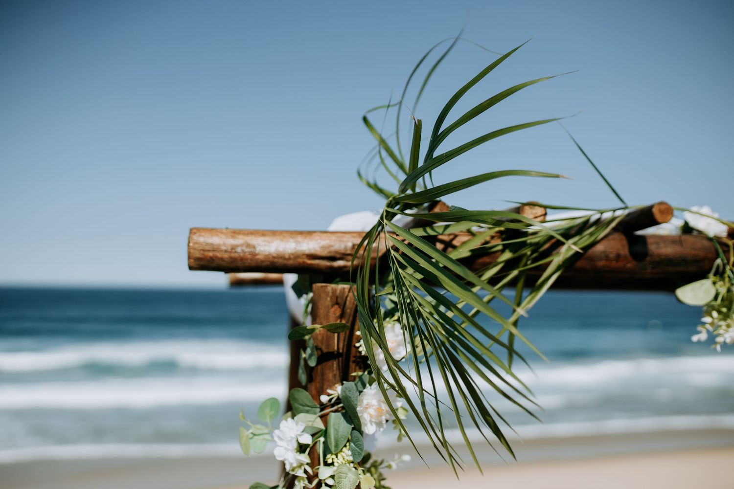 Tropical wedding ceremony setup with palm leaves and natural wood arch overlooking the ocean waves.