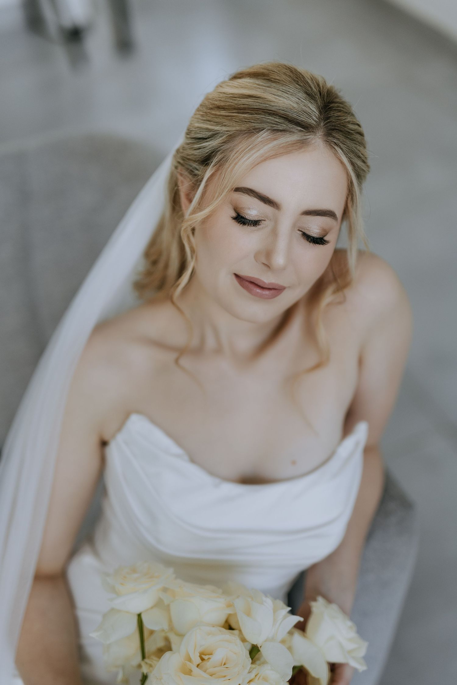 A bride in a white wedding dress holds a bouquet of cream roses while looking down with closed eyes.
