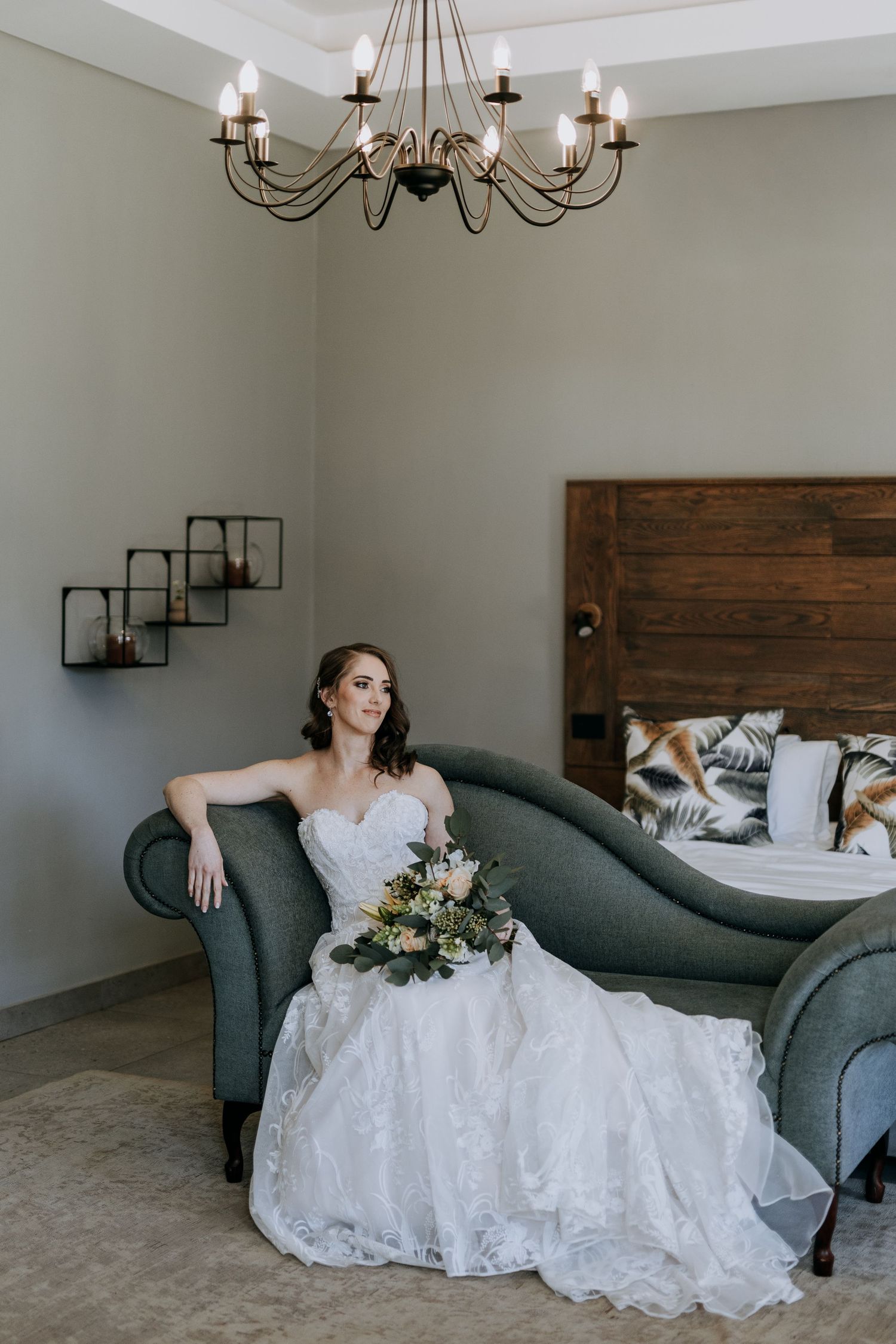 A person in a white lace wedding gown reclines gracefully on a dark gray chaise lounge beneath an elegant chandelier.