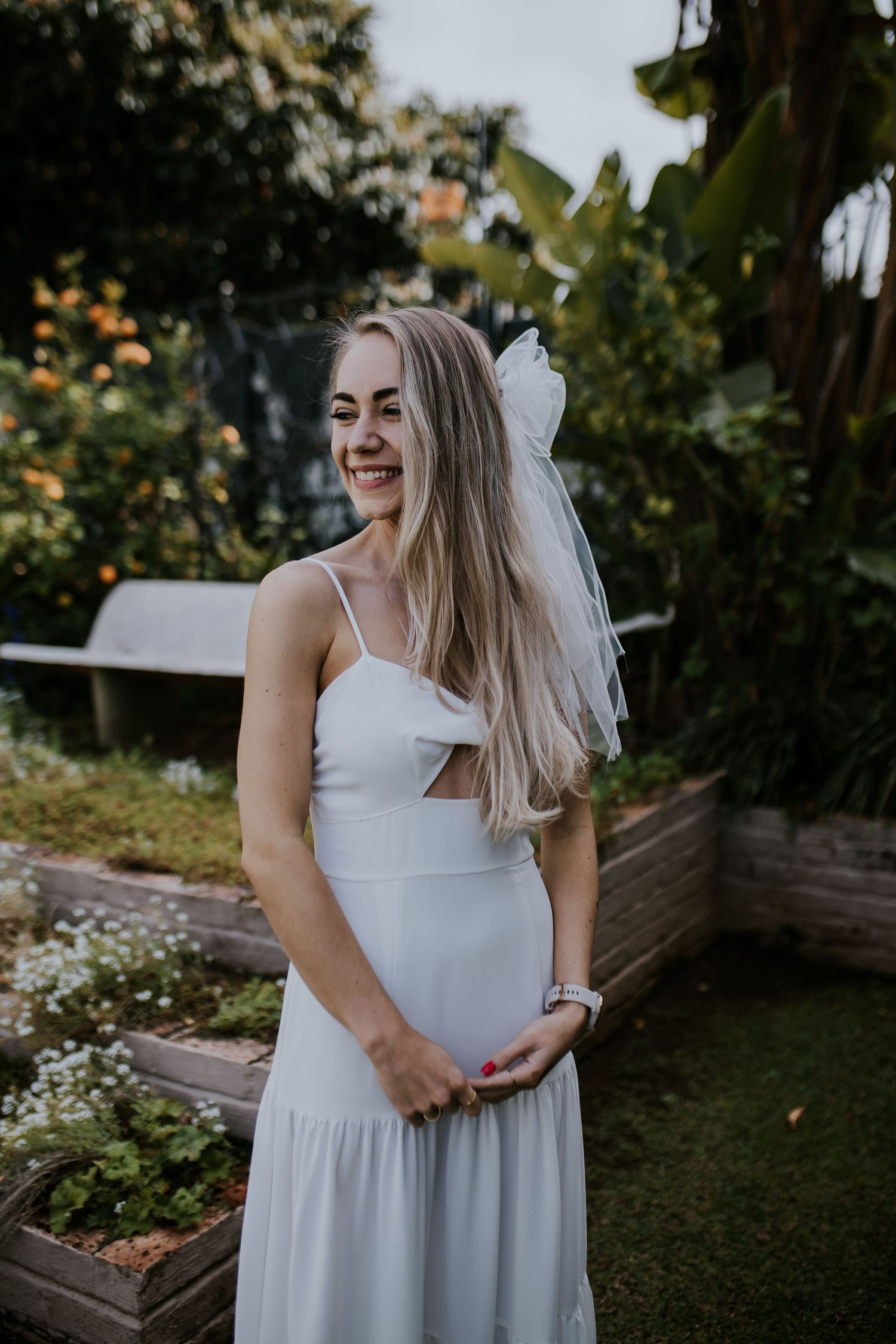 A woman in a white summer dress stands in a garden setting with blooming flowers and greenery in the background.
