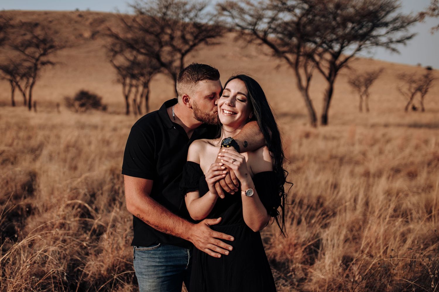 Sweet romantic couple embraces in golden sunset light on African savanna with acacia trees in background.