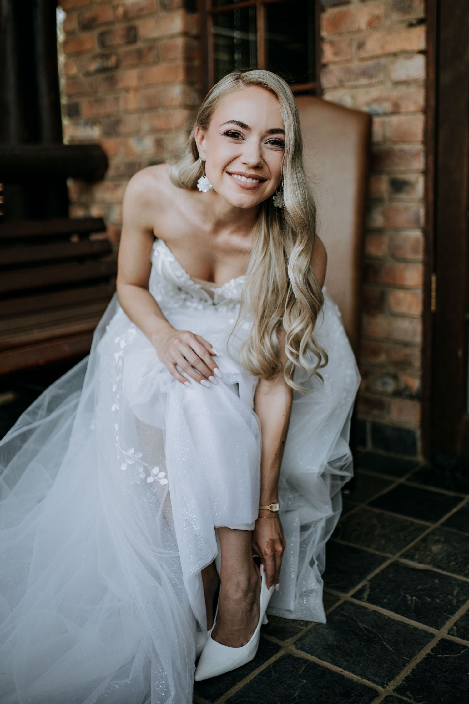 A bride in a white strapless wedding gown and heels adjusts her shoe while smiling on a brick porch.
