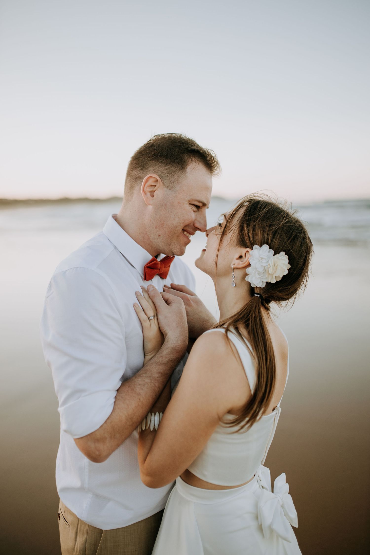 A romantic beach wedding portrait of a couple embracing at sunset, with the bride wearing a white dress and floral hair accessory.