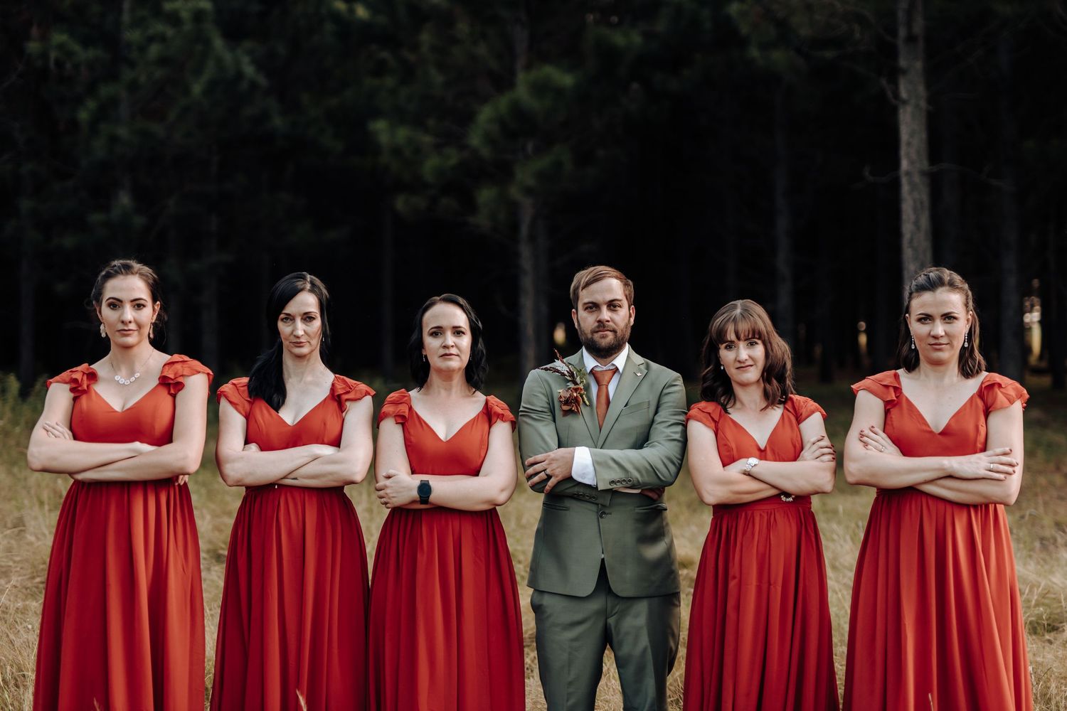 Wedding party poses together outdoors wearing red bridesmaid dresses and green suits in a forest setting.