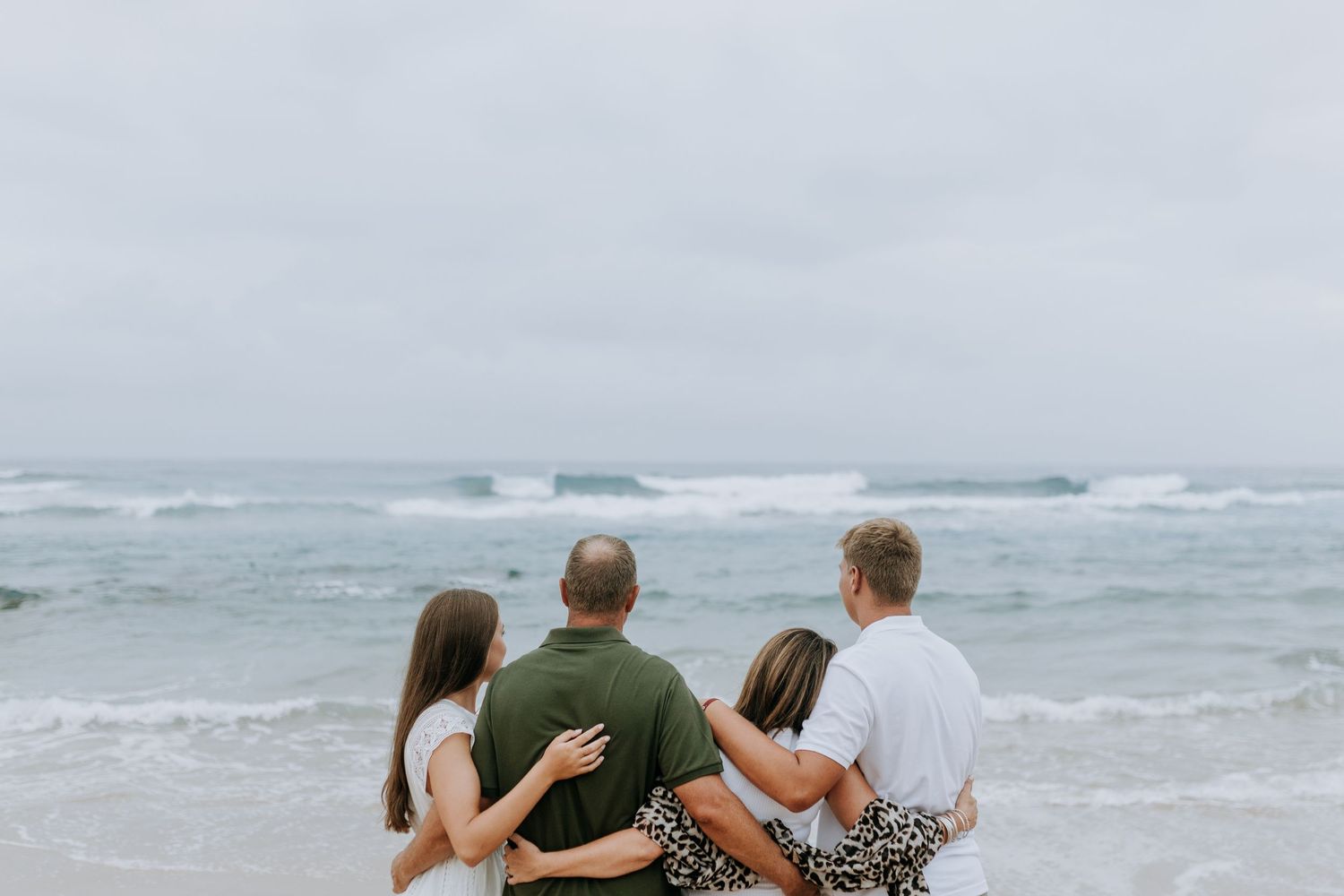 Family sits on beach watching waves crash against shoreline under cloudy skies at the ocean.