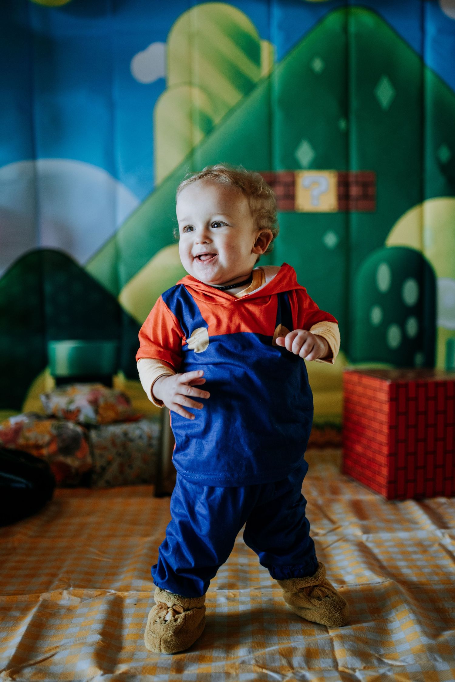 Toddler in blue overalls and red shirt crouches on patterned floor in colorful indoor playroom setting.