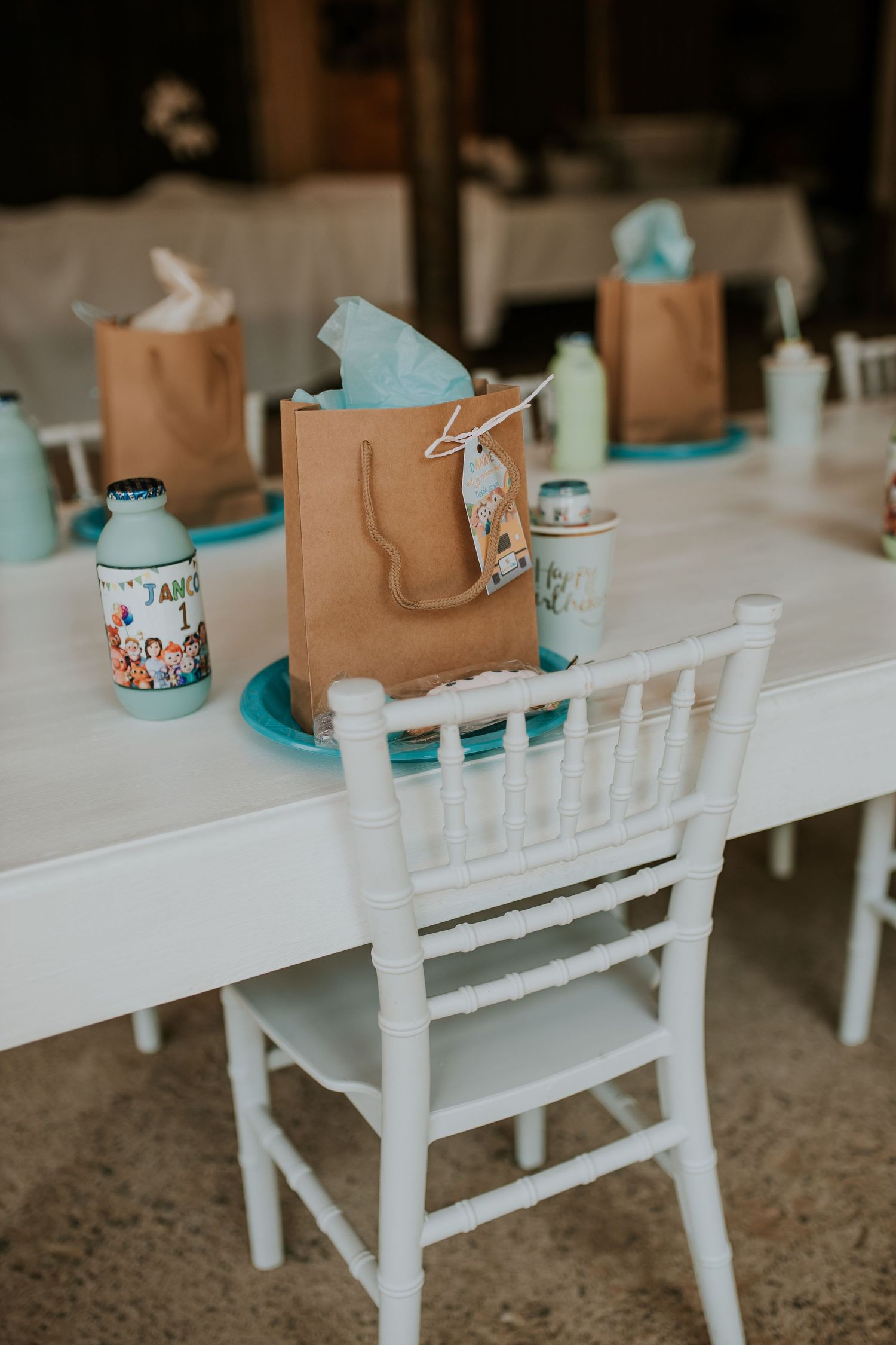 Brown paper gift bags and turquoise party decorations arranged on white wooden chairs in a rustic party setup.