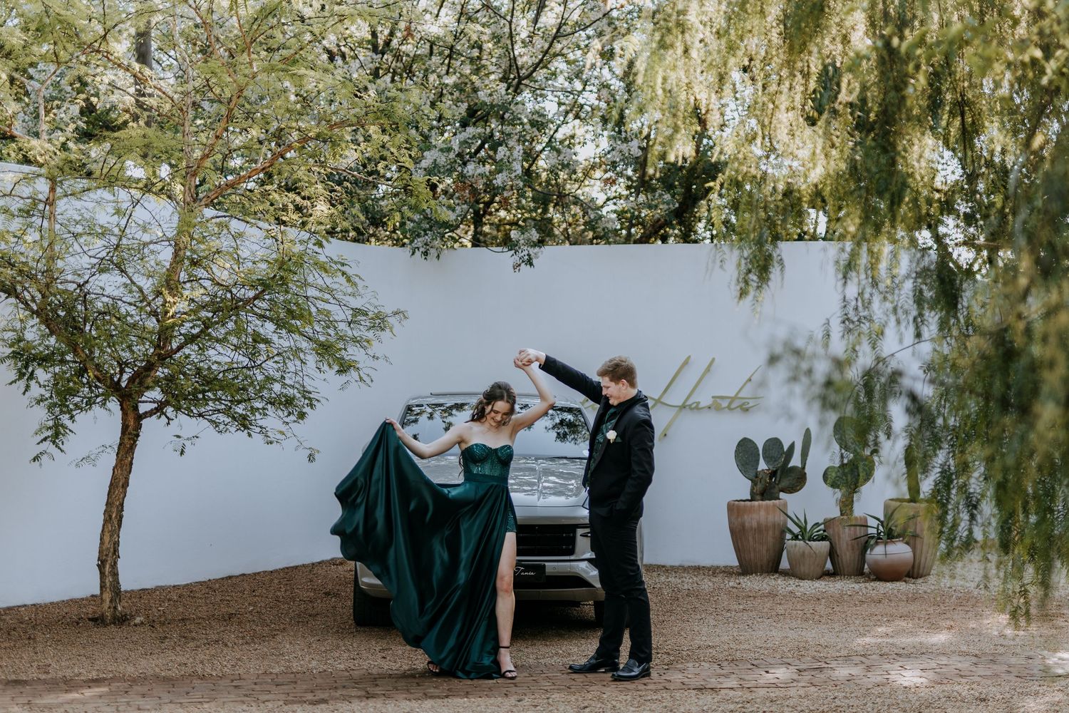 A couple shares an intimate dance moment on a beach with willow trees and chairs in the background.