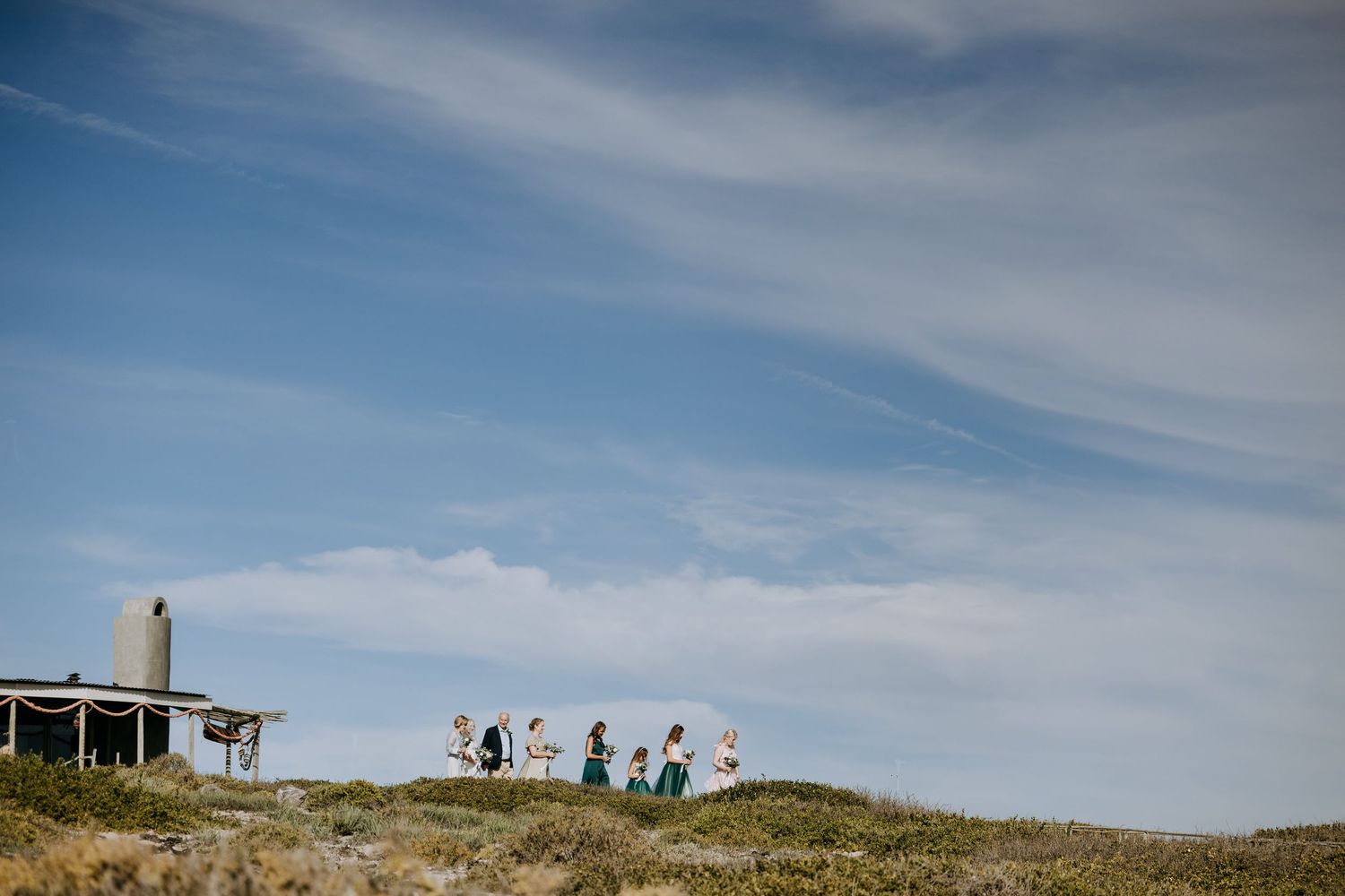 Small groups of hikers trek along the ridge of a scenic coastal path under bright blue skies and scattered clouds.