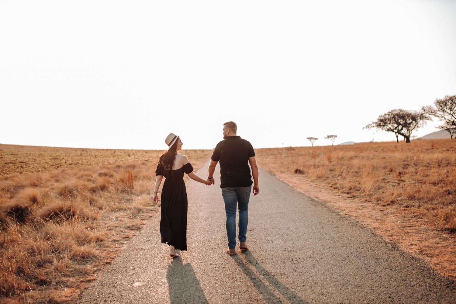 Couple holding hands walking down a dirt road in golden desert landscape at sunset creating romantic silhouettes.