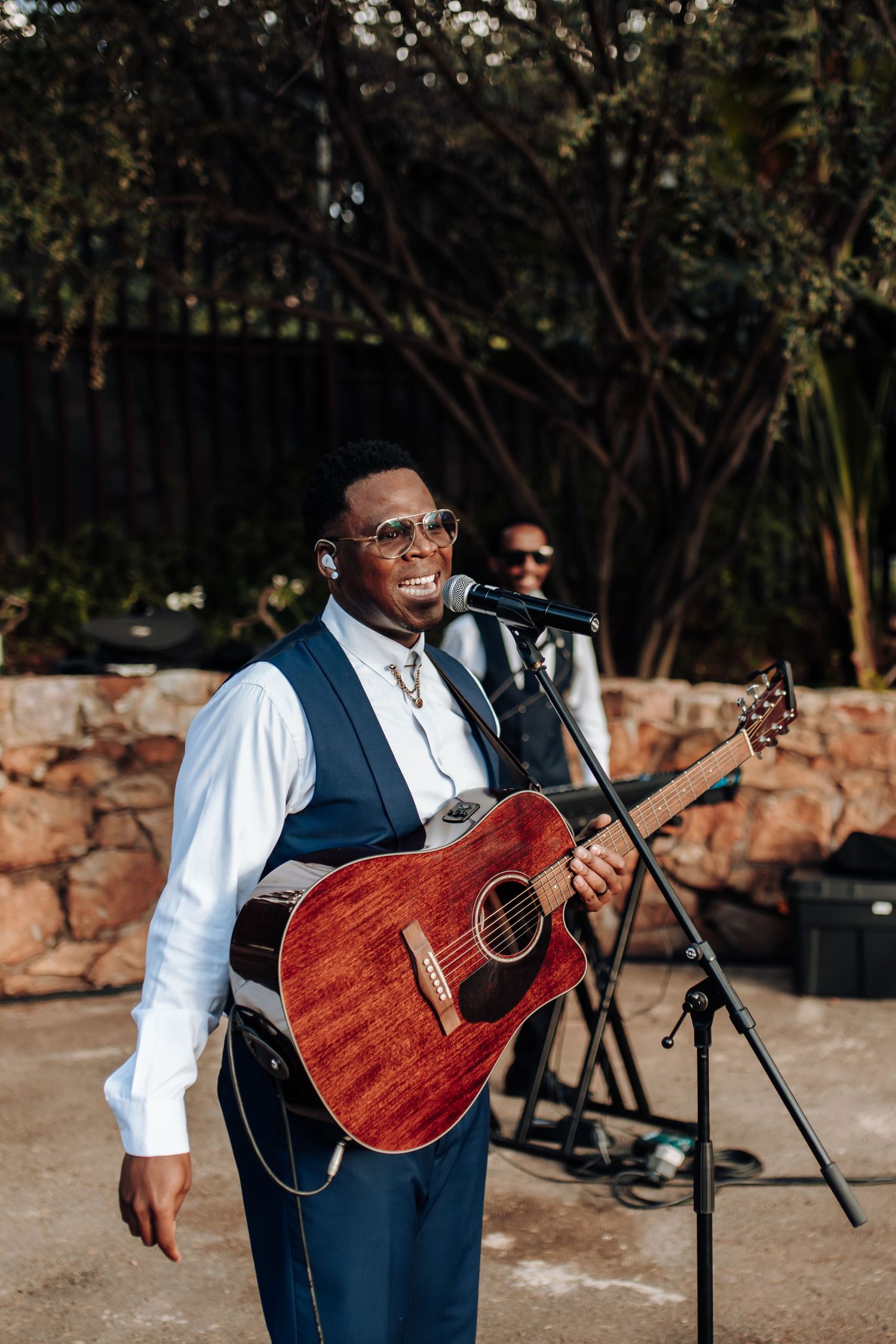 Musician in formal attire plays acoustic guitar during outdoor performance at stone wall venue in natural lighting.