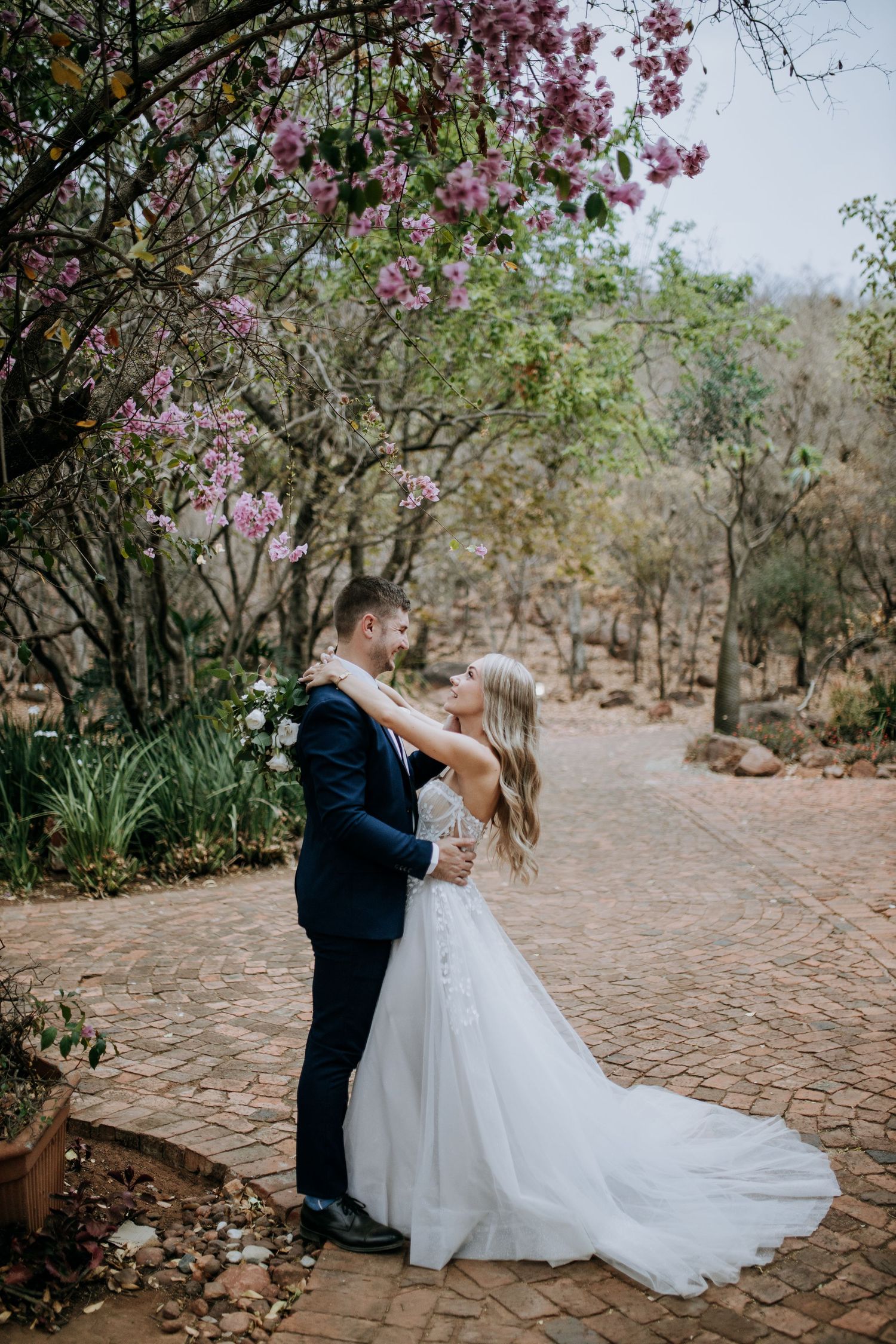 Romantic wedding couple embraces under blooming cherry blossom branches along a cobblestone path.