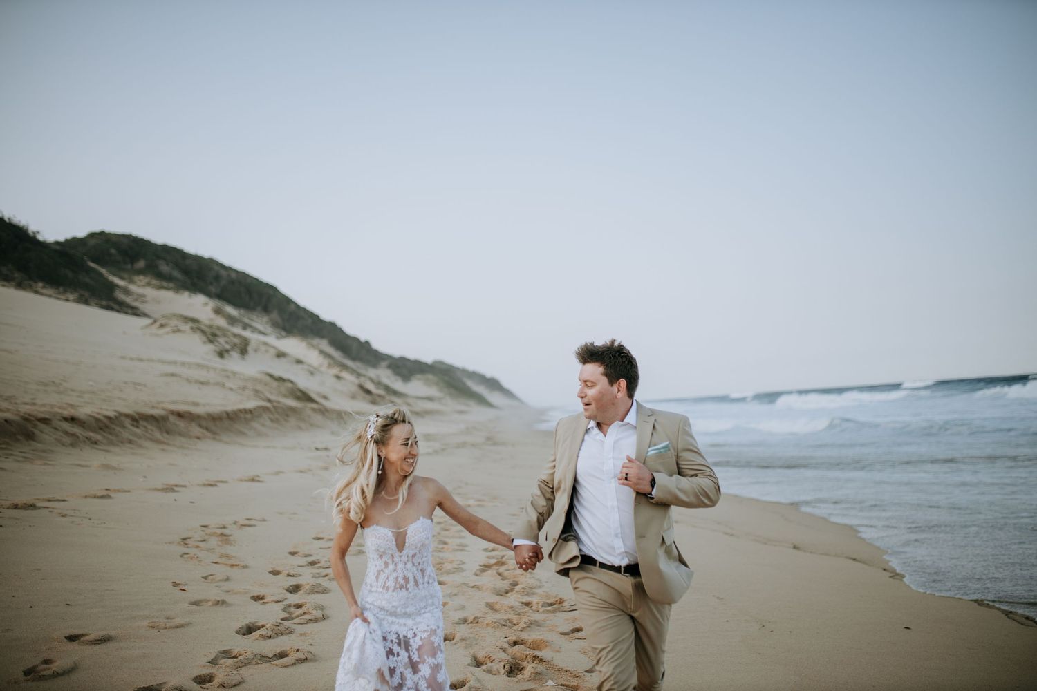 A couple walks along a misty beach together holding hands on a cloudy day with rolling waves in the background.