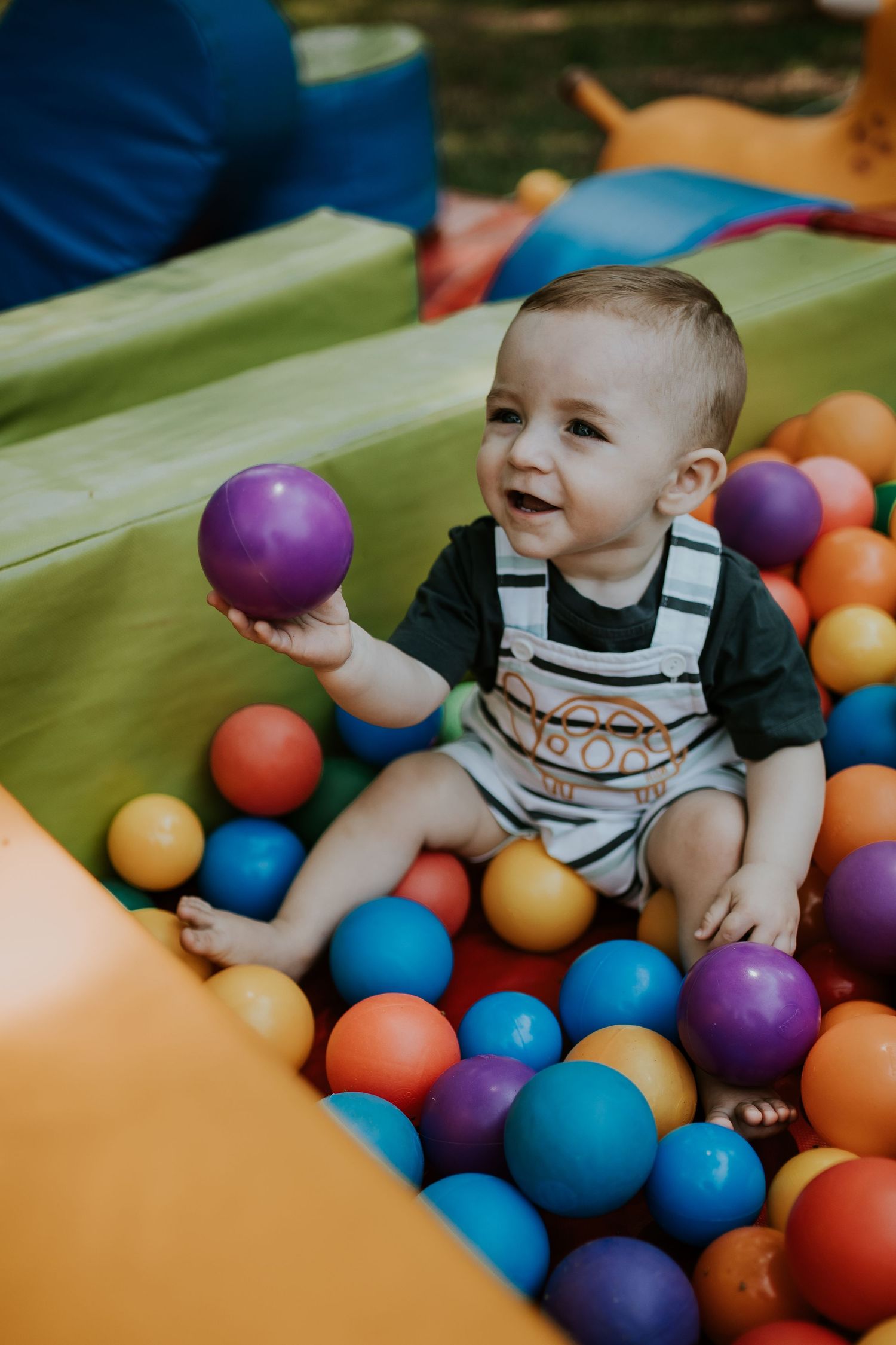 Infant in striped overalls plays joyfully in a colorful ball pit with blue, orange, and purple plastic balls.