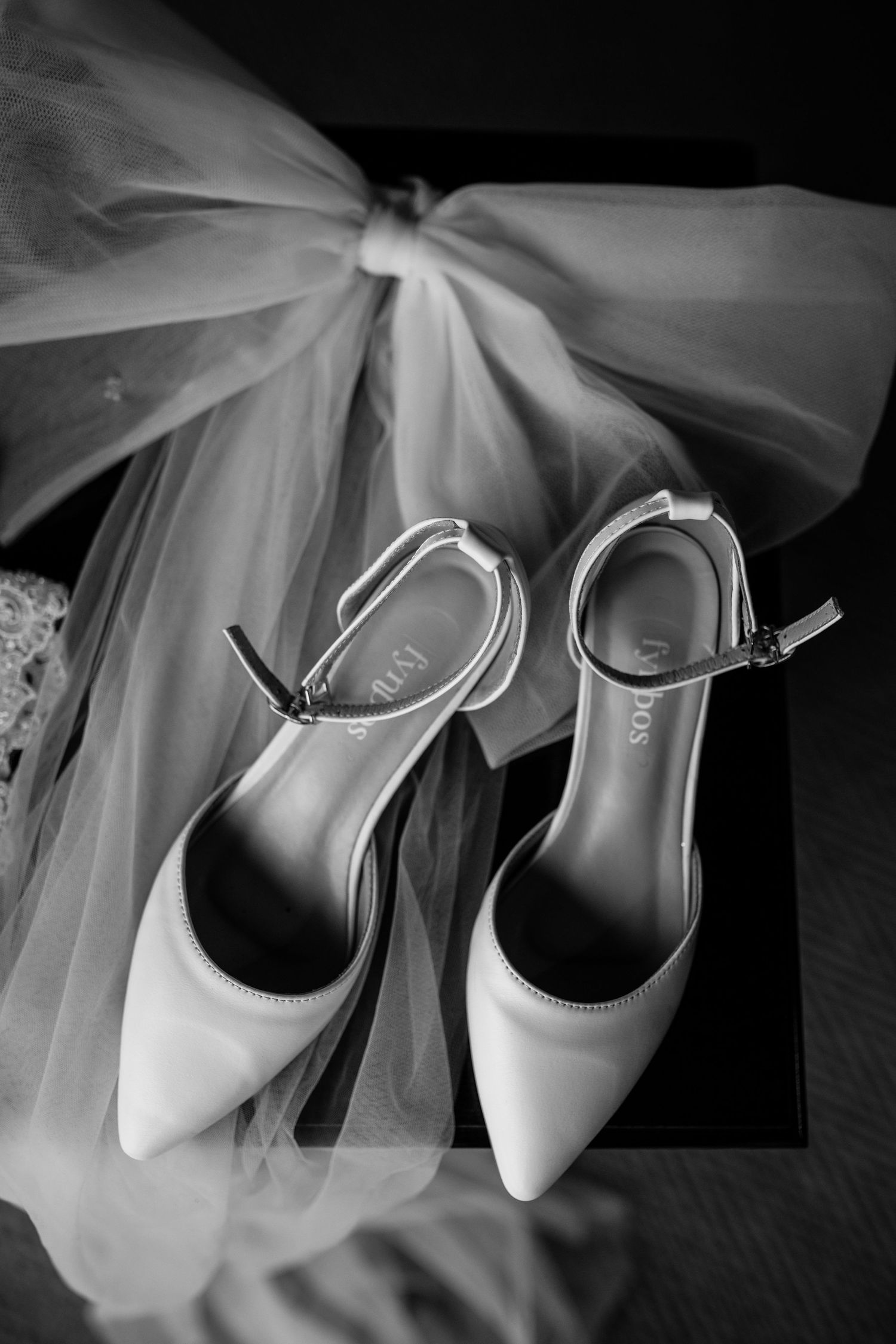 Elegant white bridal shoes with ankle straps displayed alongside flowing tulle veil in black and white photography.