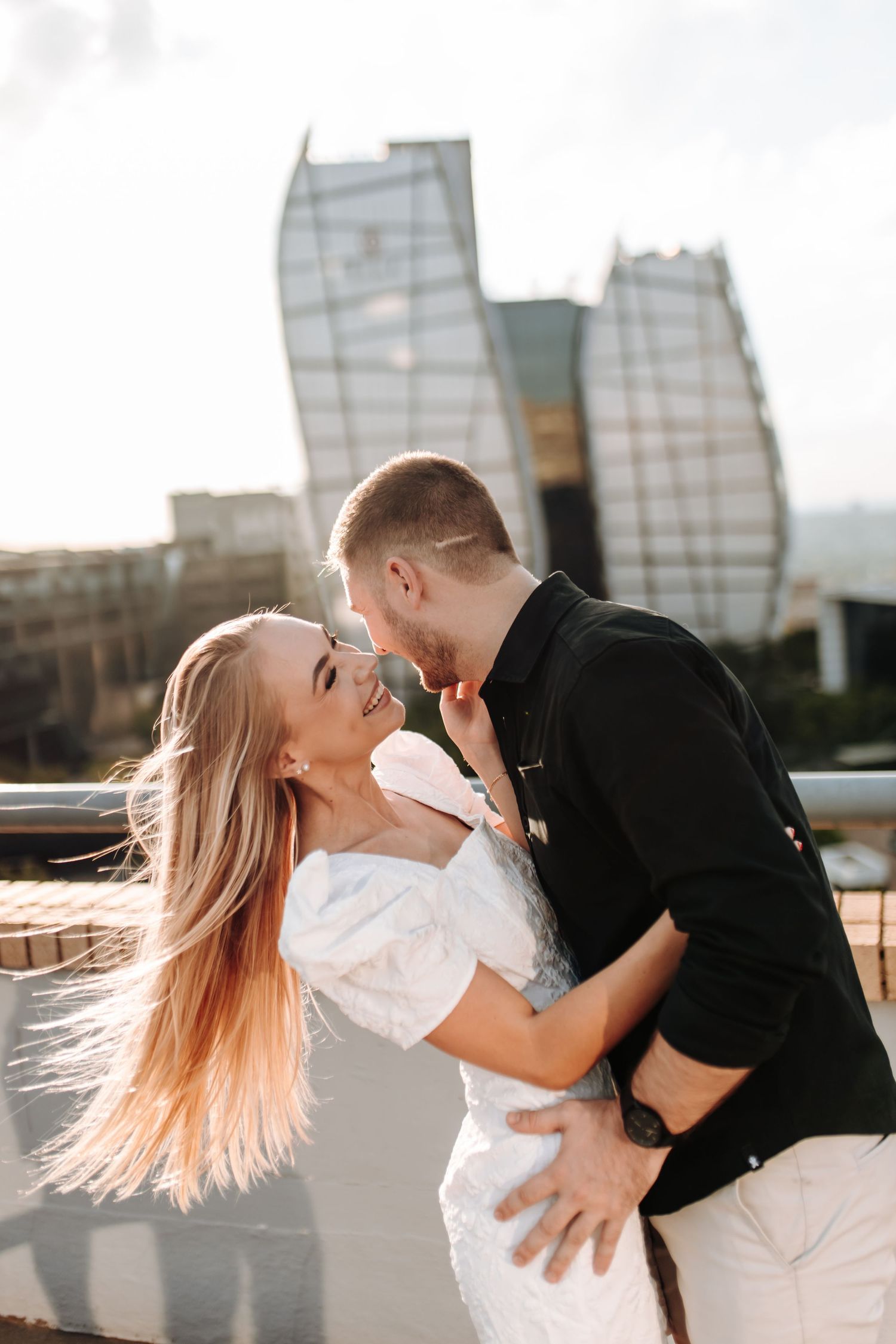 Romantic couple embraces and kisses against urban backdrop with modern glass buildings at golden hour.